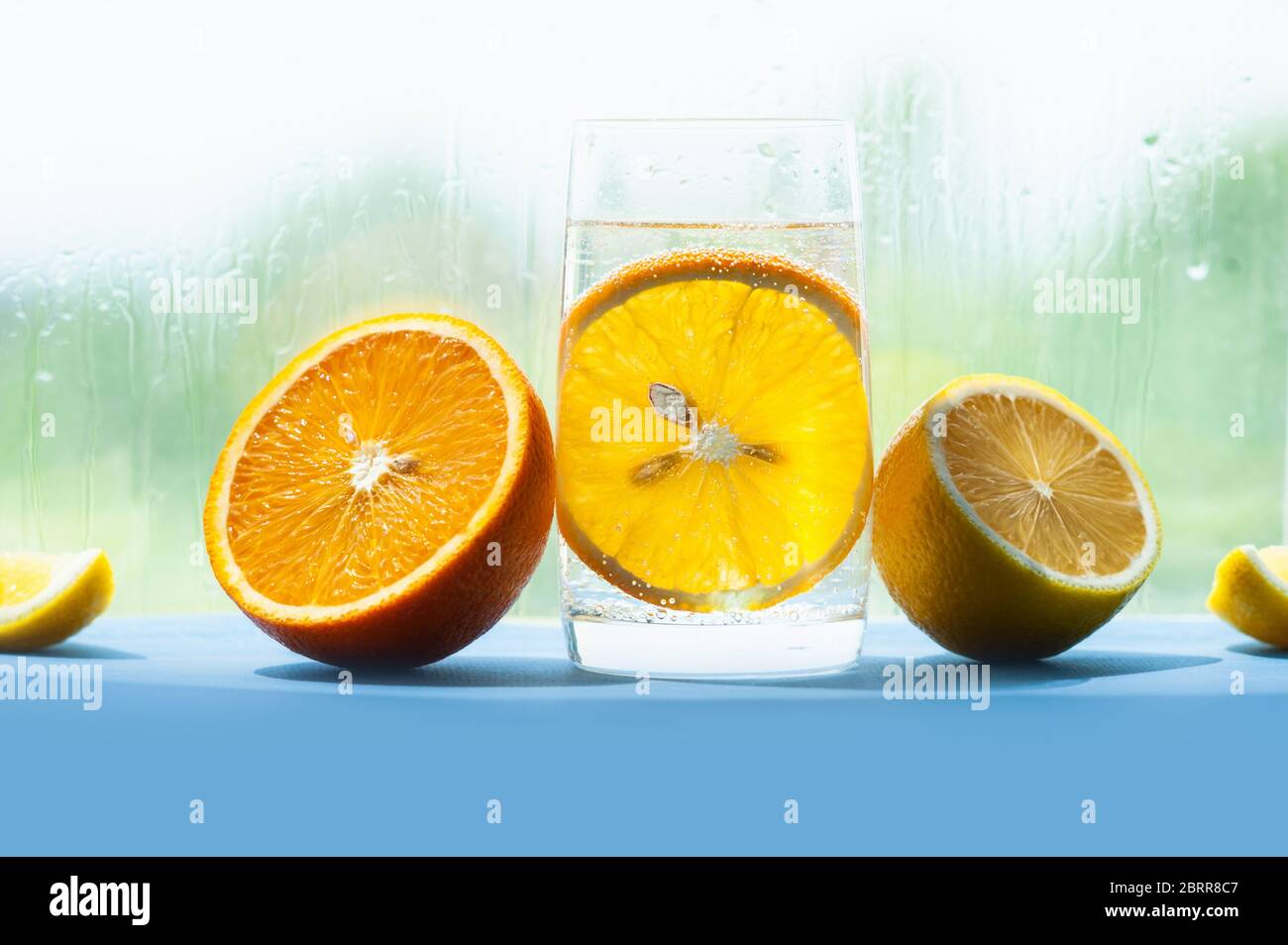 Transparentes Glas mit gasförmigem Wasser und Blasen und einer runden Scheibe Orange innen, halb saftig und reif Orange und Zitrone daneben, Glas mit Tropfen Stockfoto