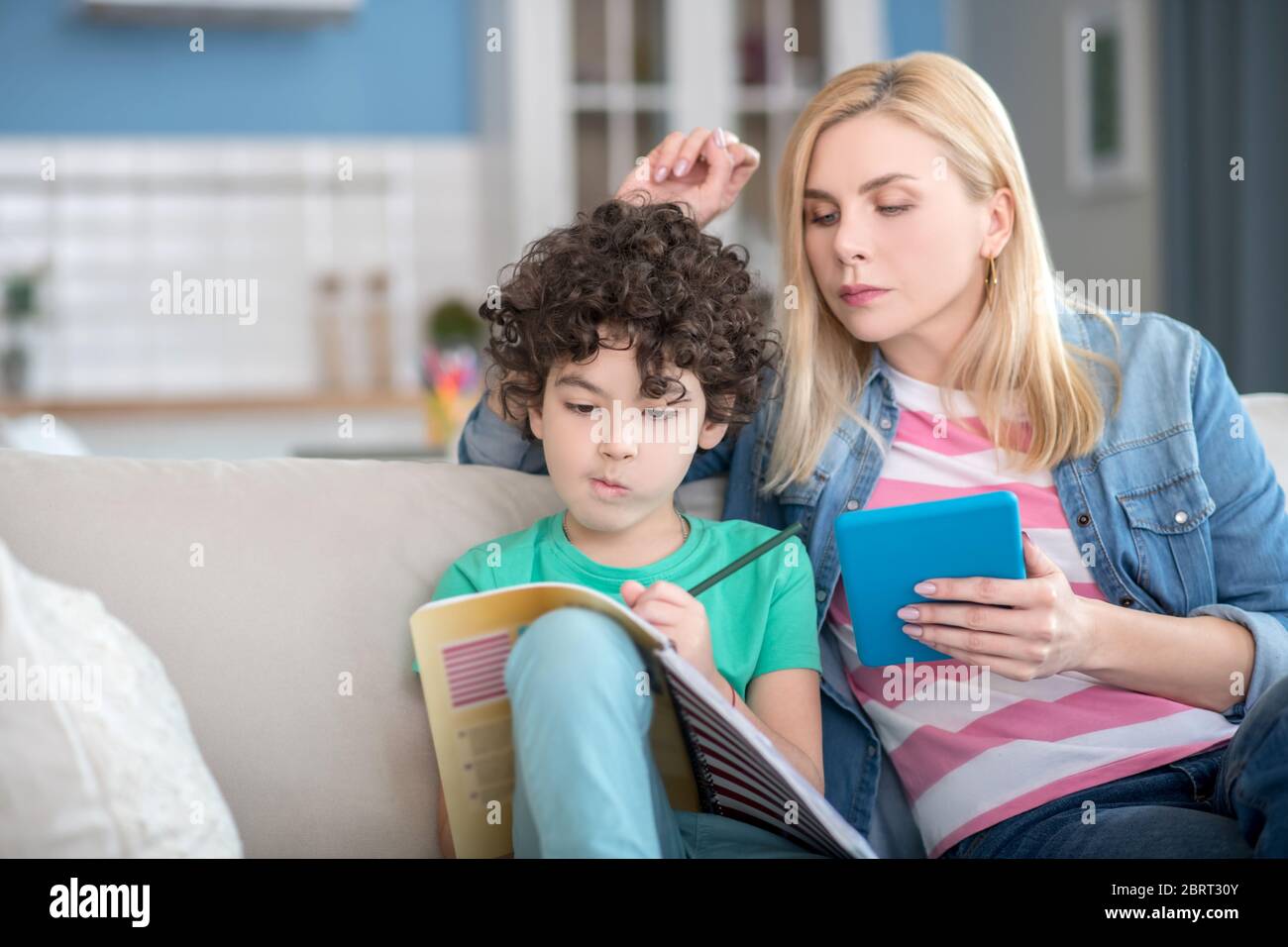 Blonde Frau hält Tablette, was lockig Jungen in seinem Notizbuch schreiben zu überprüfen Stockfoto