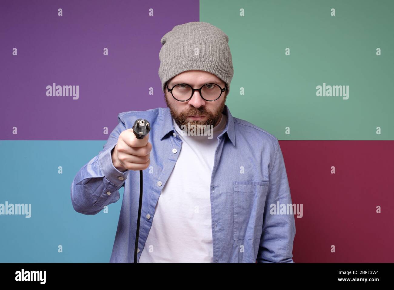 Wütender bärtiger Mann, in runden Gläsern und einem Hut, hält in den Händen einen elektrischen Stecker mit einer Schnur. Isoliert auf einem bunten Hintergrund. Stockfoto