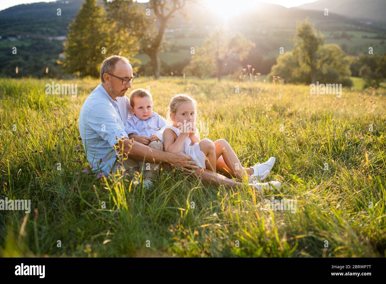 Junger Vater mit zwei kleinen Kindern, die bei Sonnenuntergang auf einer Wiese im Freien sitzen. Stockfoto