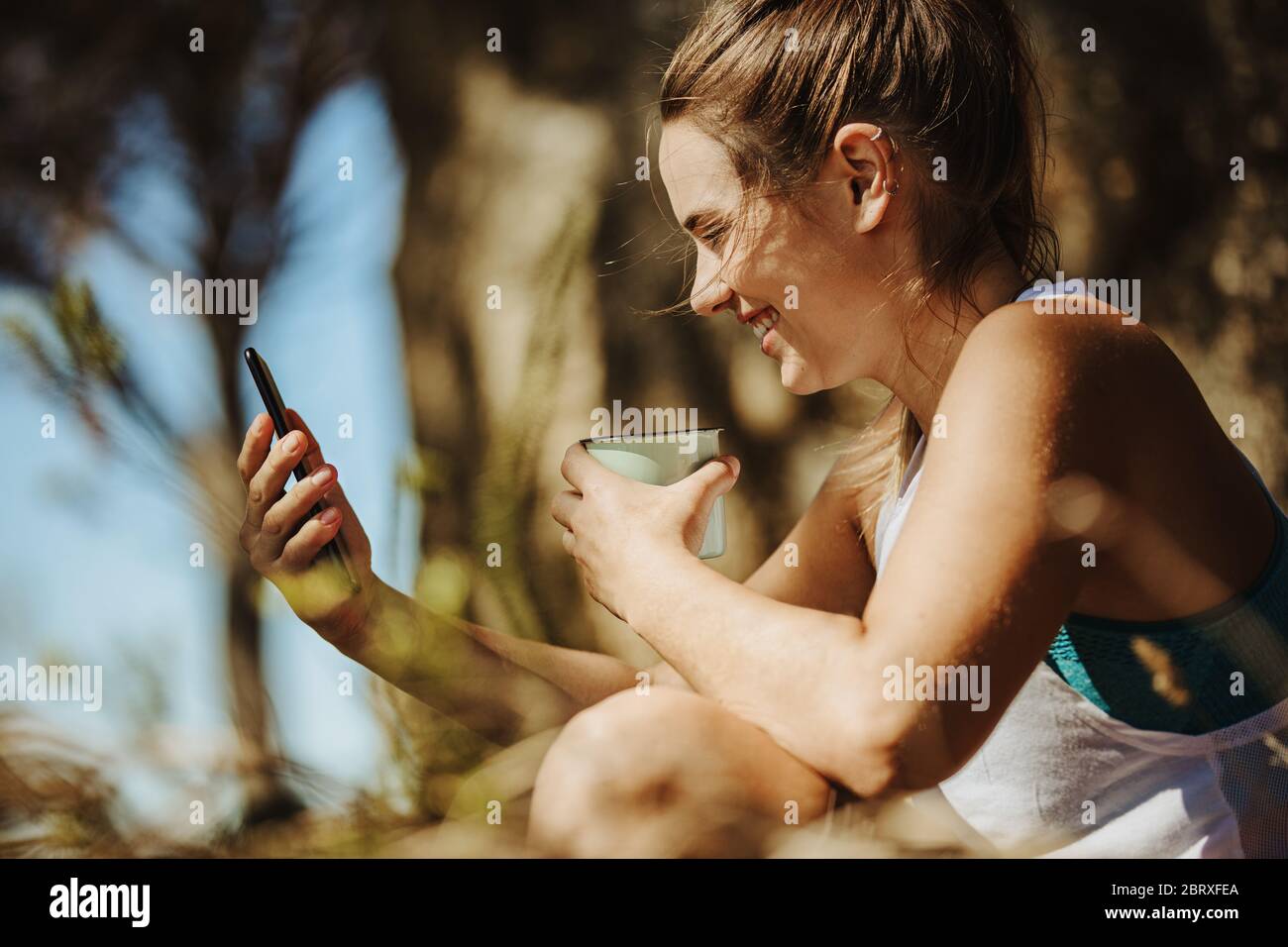 Lächelnde junge Frau, die ihr Telefon anschaut und während des Trekkings im Freien Kaffee trinkt. Sportliche Frau, die beim Wandern eine Pause einlegen. Stockfoto