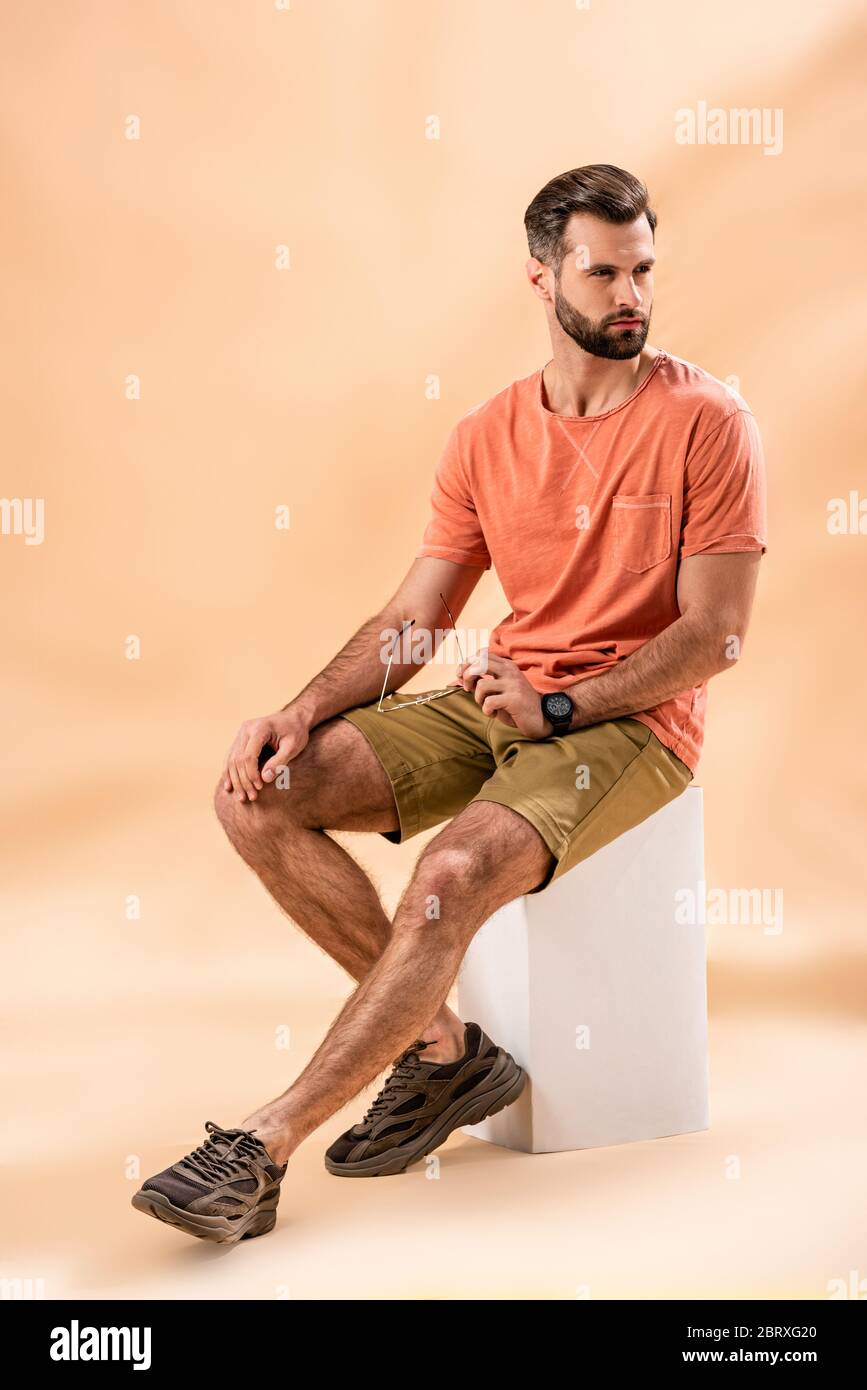 Stilvoller junger Mann in Shorts und Sommer-T-Shirt mit Sonnenbrille sitzt auf weißem Würfel auf beige Stockfoto