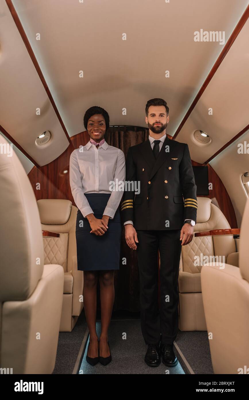 Gutaussehende, selbstbewusste Pilot und lächelnde afroamerikanische Stewardess Blick auf Kamera in Privatflugzeug Stockfoto