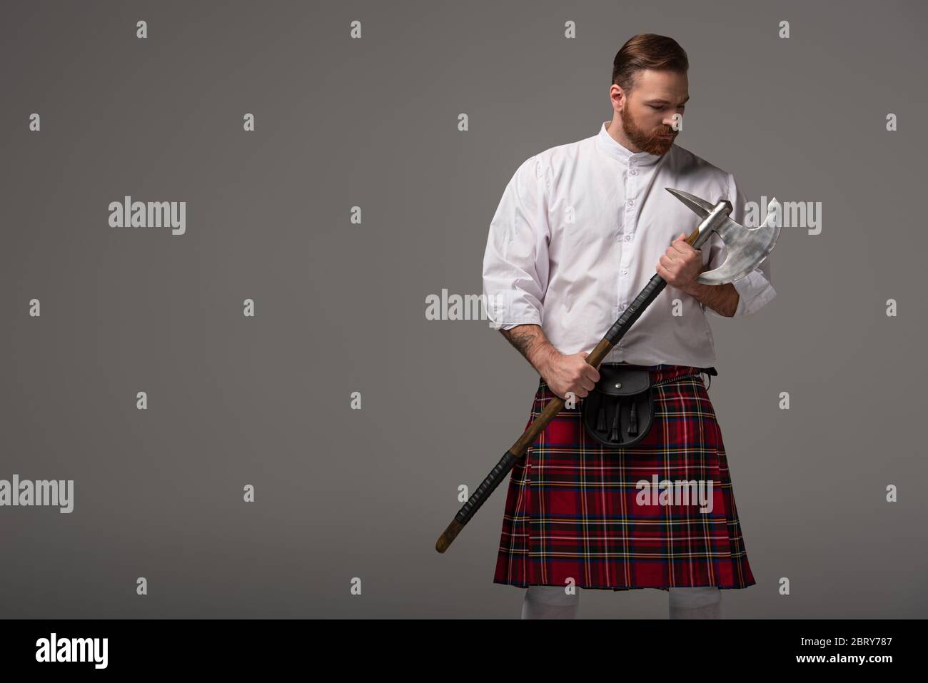Schottischer Rotschopf-Mann in rotem Kilt mit Schlachtaxt auf grauem Hintergrund Stockfoto