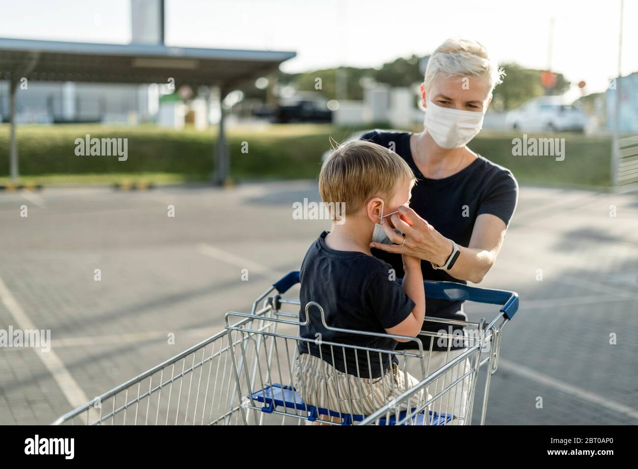 Mutter hilft ihrem Sohn, der in einem Einkaufswagen sitzt, eine Schutzmaske zu tragen, bevor er in einen Laden kommt. Stockfoto