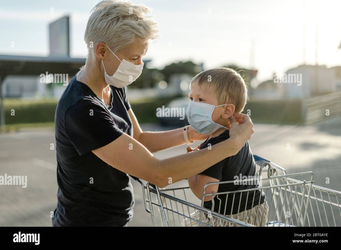 Mutter hilft ihrem Sohn, der in einem Einkaufswagen sitzt, eine Schutzmaske zu tragen, bevor er in einen Laden kommt. Stockfoto
