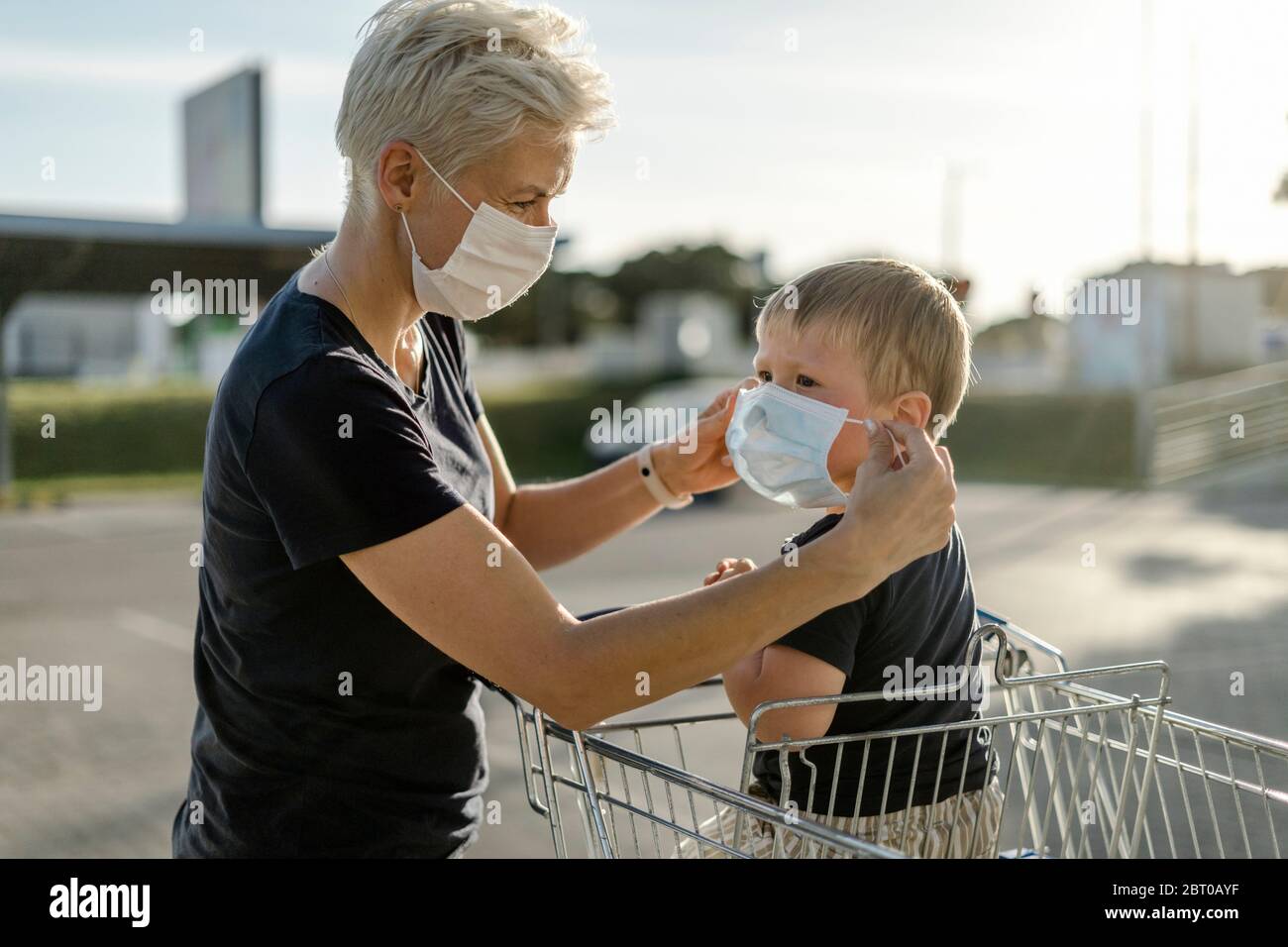Mutter hilft ihrem Sohn, der in einem Einkaufswagen sitzt, eine Schutzmaske zu tragen, bevor er in einen Laden kommt. Stockfoto