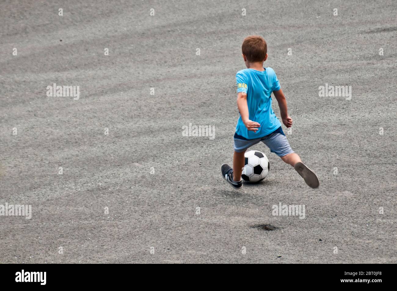 Kind in blauem Hemd und Shorts treten einen Fußball auf der Straße Stockfoto