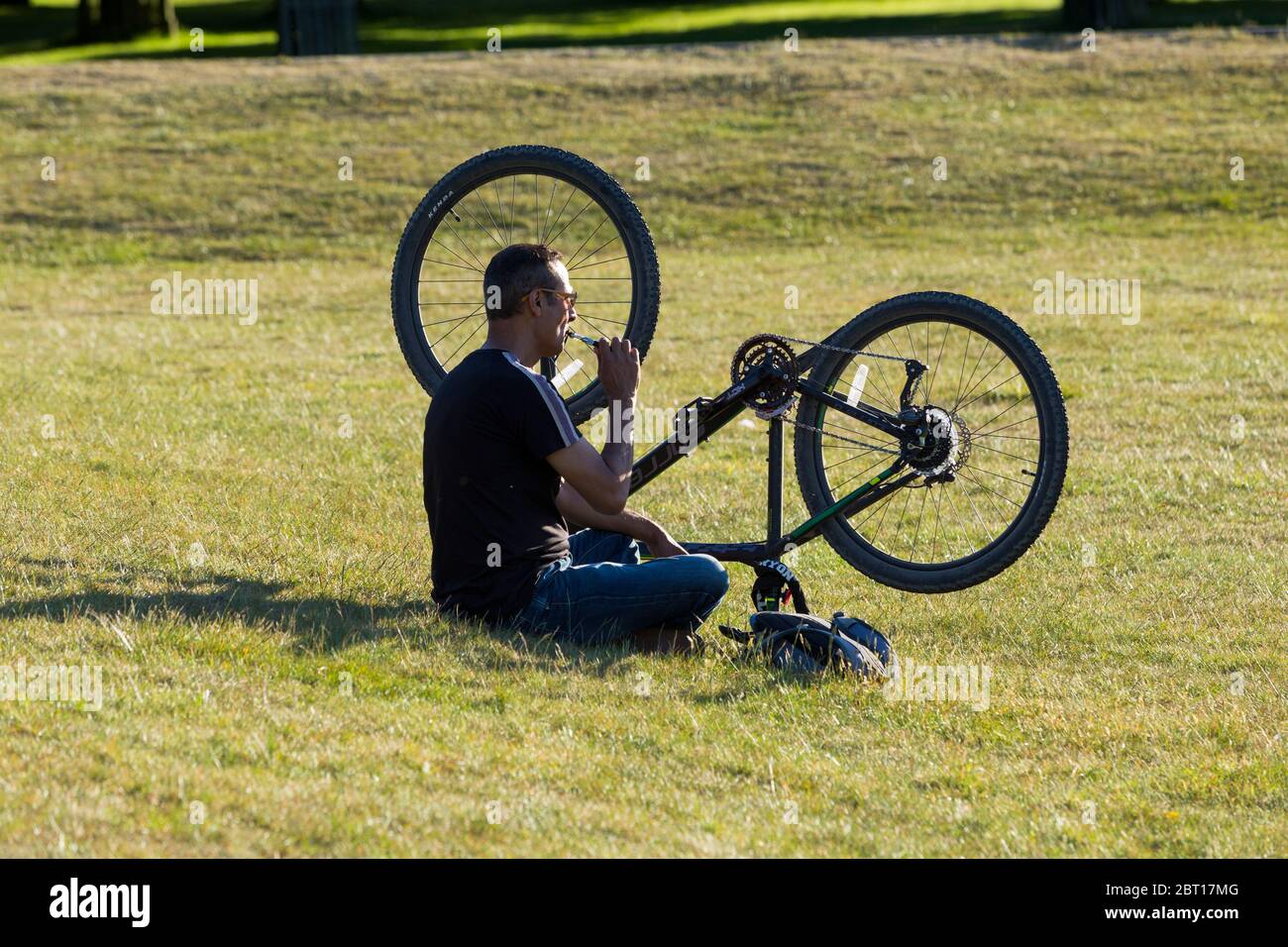 Ein Mann mit Radler entspannt sich in einem öffentlichen Park auf dem Gras und genießt den sonnigen Sonnentag mit seinem Fahrrad auf den Kopf gestellt. Er raucht ein nicht-Tabakgerät, wie eine Vaping-Zigarette London Middlesex UK (119) Stockfoto