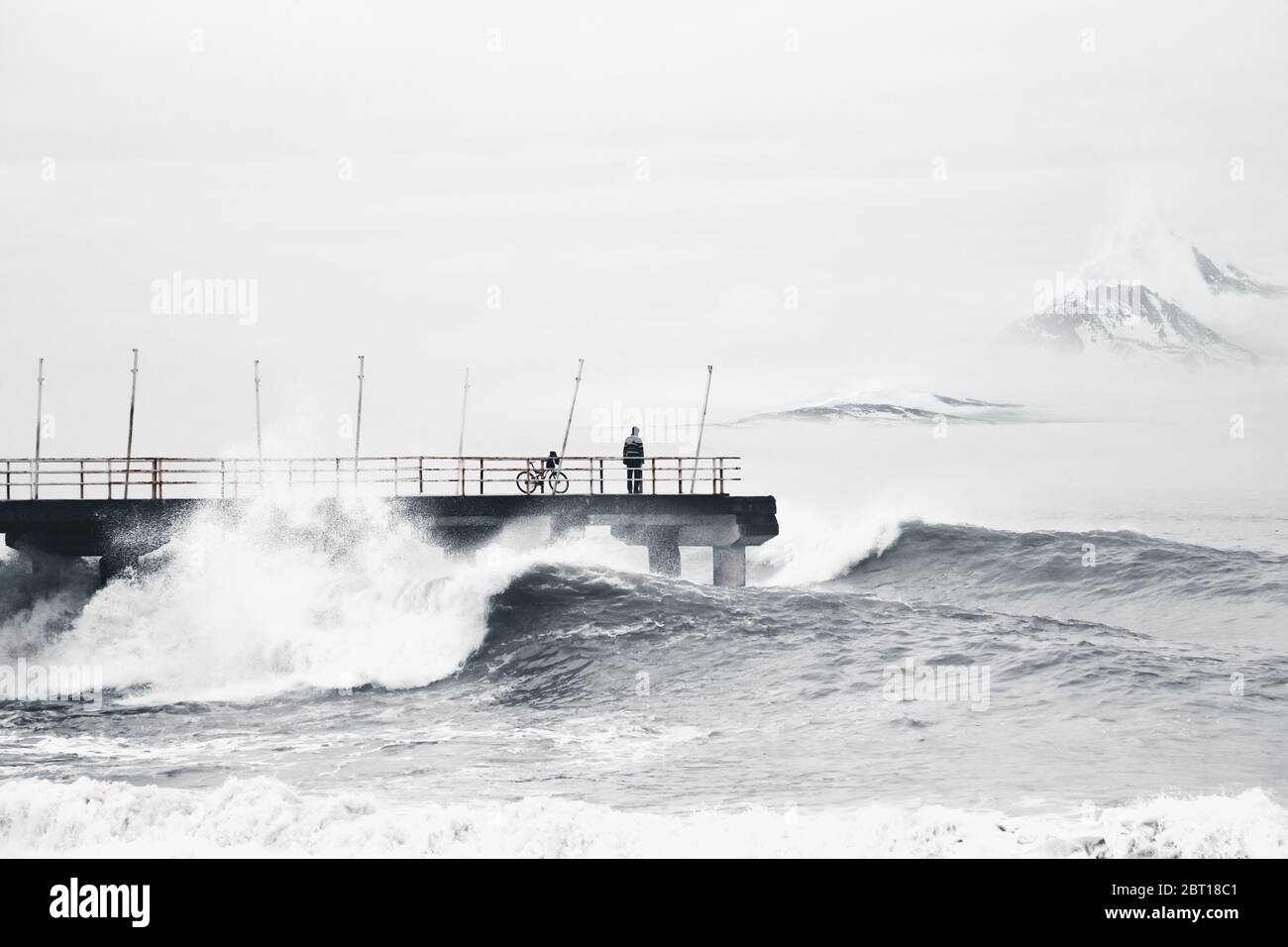 Ein Radfahrer beobachtet von der Pier aus die riesigen Wellen im Sturm. Im Nebel verschneite Alpen am Horizont. Der Mann ist mutig. Stockfoto