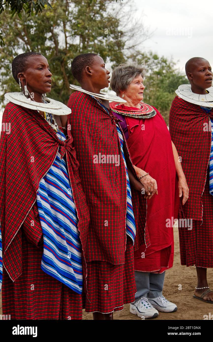 Frauen singen, drei Maasai, kaukasischer Besucher, Dorf; indigene Bevölkerung; traditionelle rote Kleidung; Perlen Doppel Hals Ringe, große Ohrringe, Loch in Stockfoto