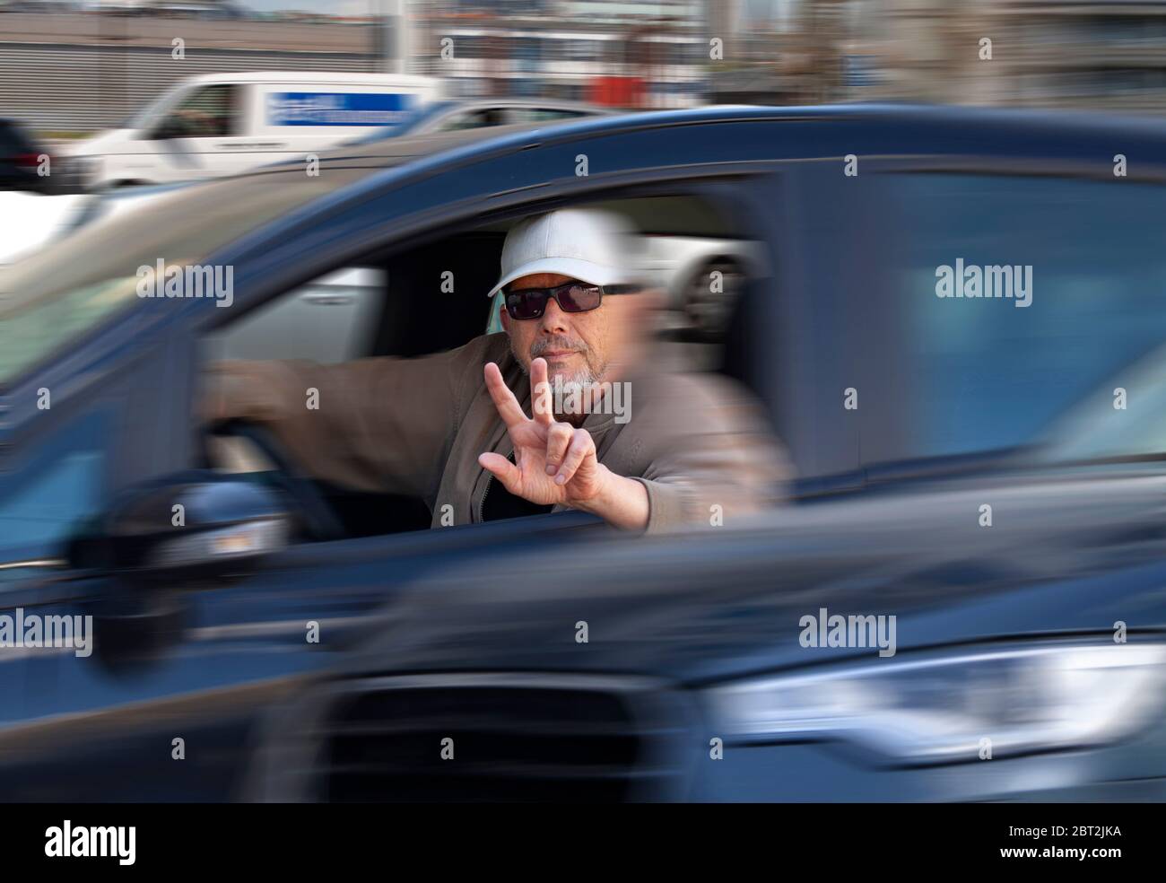 Männlicher Fahrer mit Sonnenbrille und Mütze während der Fahrt im schweren Verkehr das V-Zeichen für das Victory Peace Symbol mit den Fingern gestikuliert. Teilweise Bewegungsunschärfe Stockfoto
