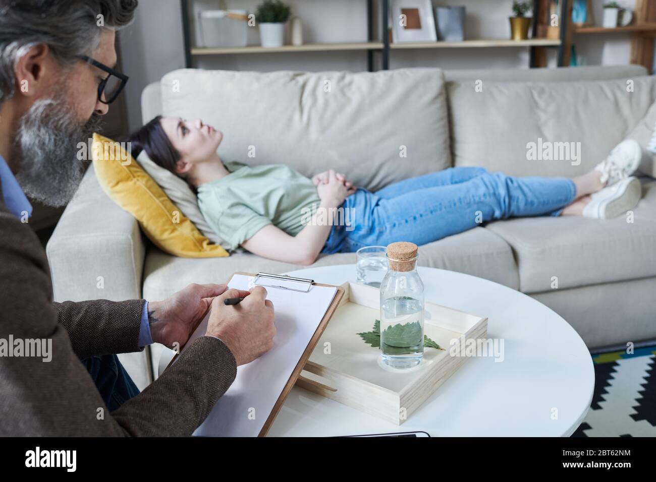 Bärtige reife männliche Berater schriftlich Probleme eines seiner Patienten liegen auf dem Sofa in der Nähe und teilen ihre Ängste oder Probleme Stockfoto