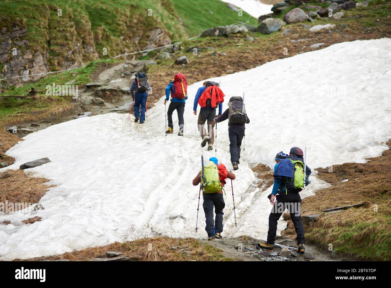 Rückansicht von Wanderern mit Rucksäcken und Wanderstöcken, die auf schneebedeckter Straße in den Bergen wandern. Gruppe von Touristen, die den Hügel klettern, während sie zusammen reisen. Konzept des Wanderns und Bergsteigens. Stockfoto