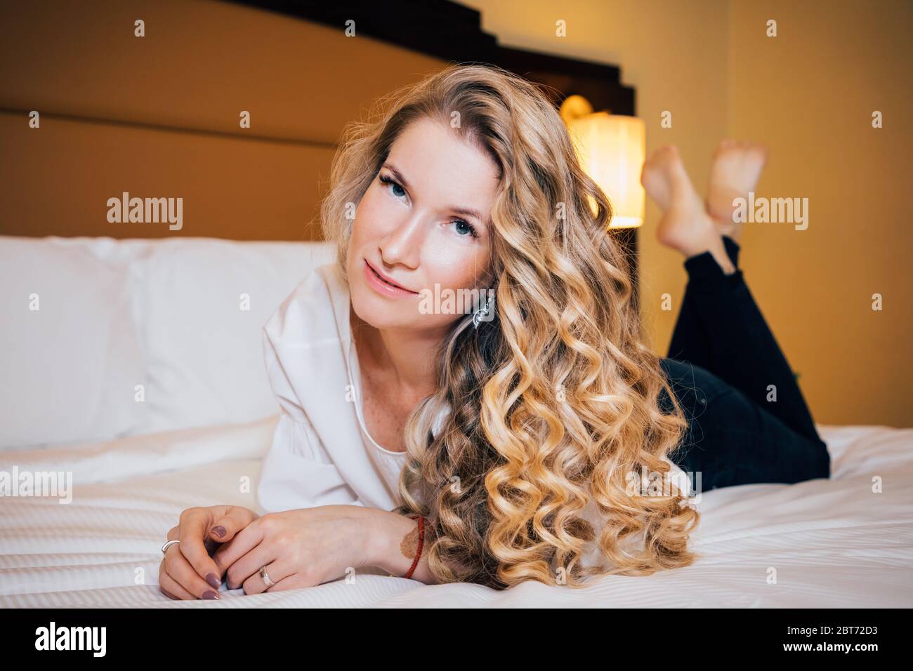 Schönes Mädchen mit langen lockigen blonden Haaren, trägt schwarze Hosen und ein helles Top, liegt auf einem Bett mit weißen Laken und Kissen. Sie schaut uns an Stockfoto