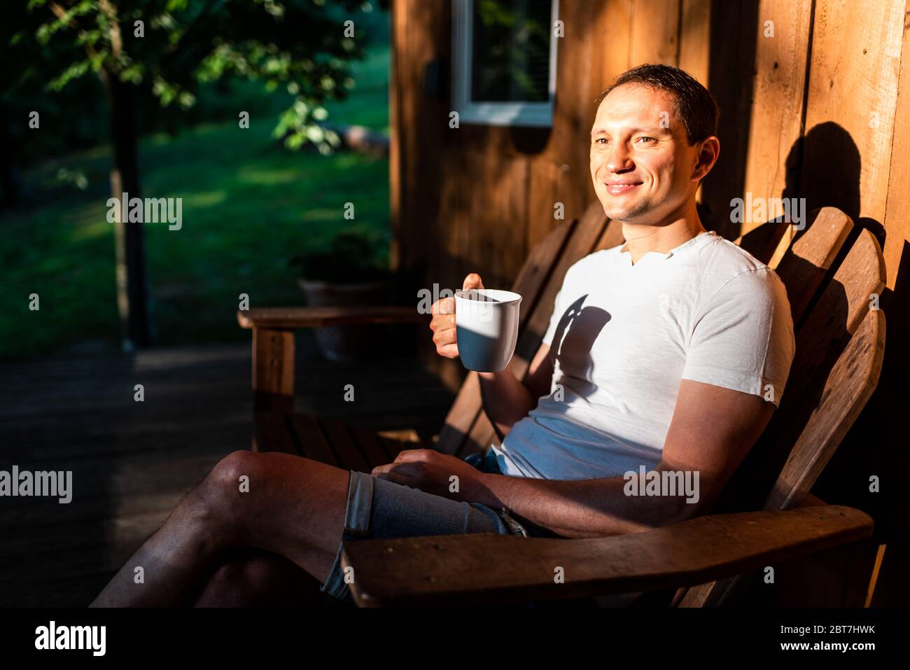 Mann glücklich sitzen entspannen auf Schaukelstuhl Lounge auf der Veranda des Hauses am Morgen Holzhütte trinken Kaffee oder Tee aus Tasse Becher Stockfoto