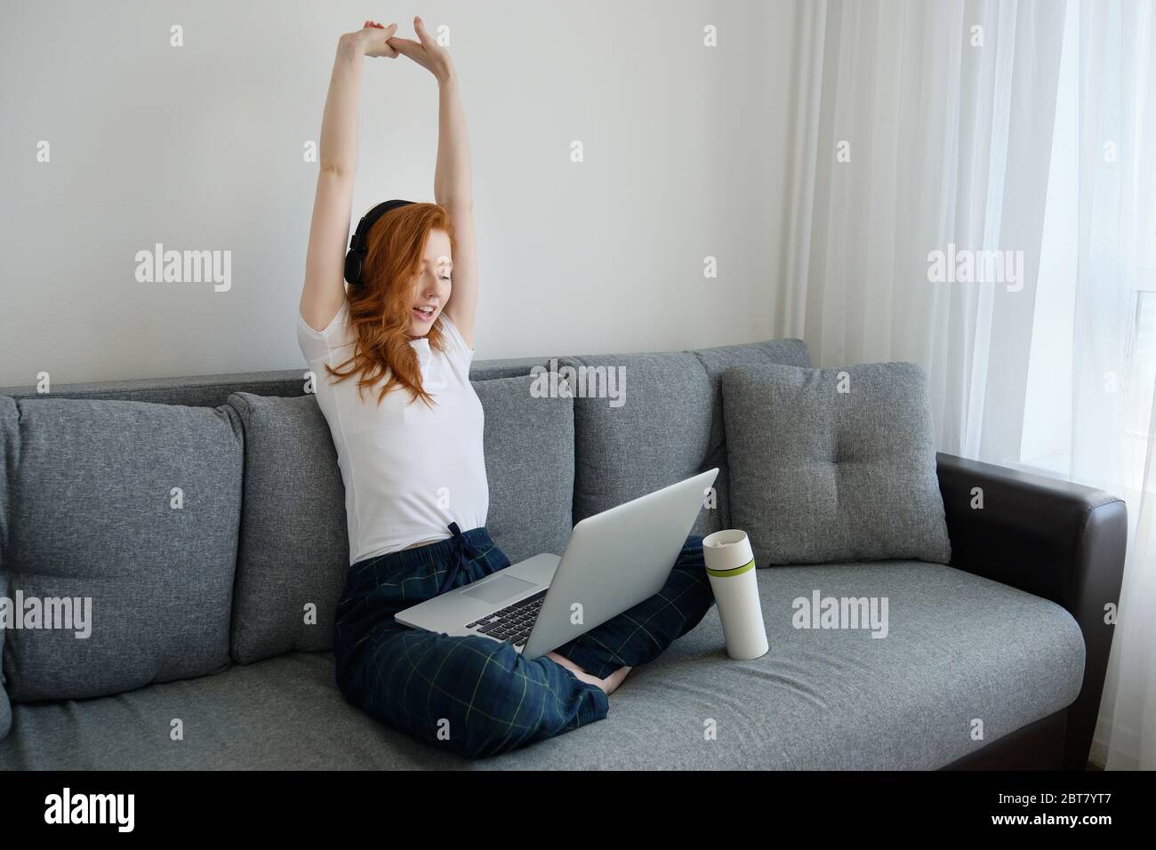 Rothaarige Mädchen sitzt auf einem Sofa in Heimkleidung mit einem Laptop auf gekreuzten Beinen und gestreckten Armen Stockfoto