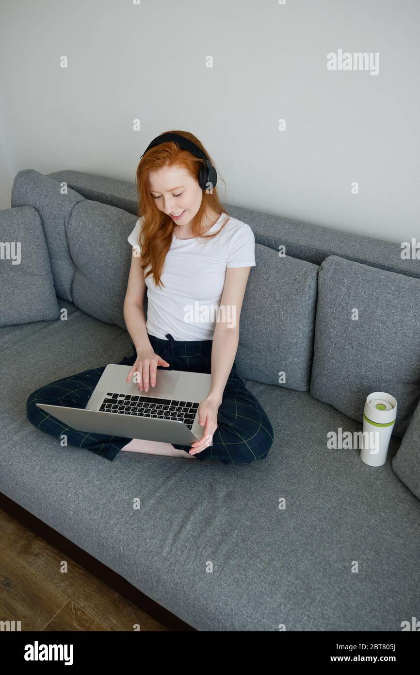 Rotschopf Mädchen sitzt auf einem Sofa in Heimkleidung und Kopfhörer mit einem Laptop auf gekreuzten Beinen, Rahmen von oben Stockfoto