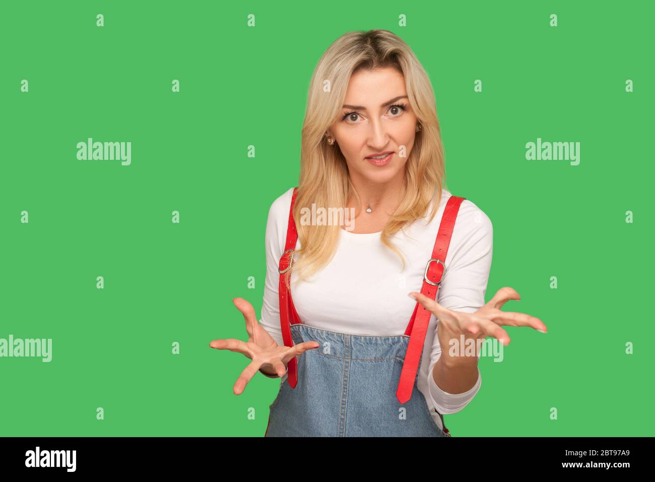 Streit, aggressive Emotionen. Porträt von gereizten Erwachsenen blonde Frau in stilvollen Denim Overalls Heben ihre Hände in Wut, klammernden Zähnen und Anschauung Stockfoto