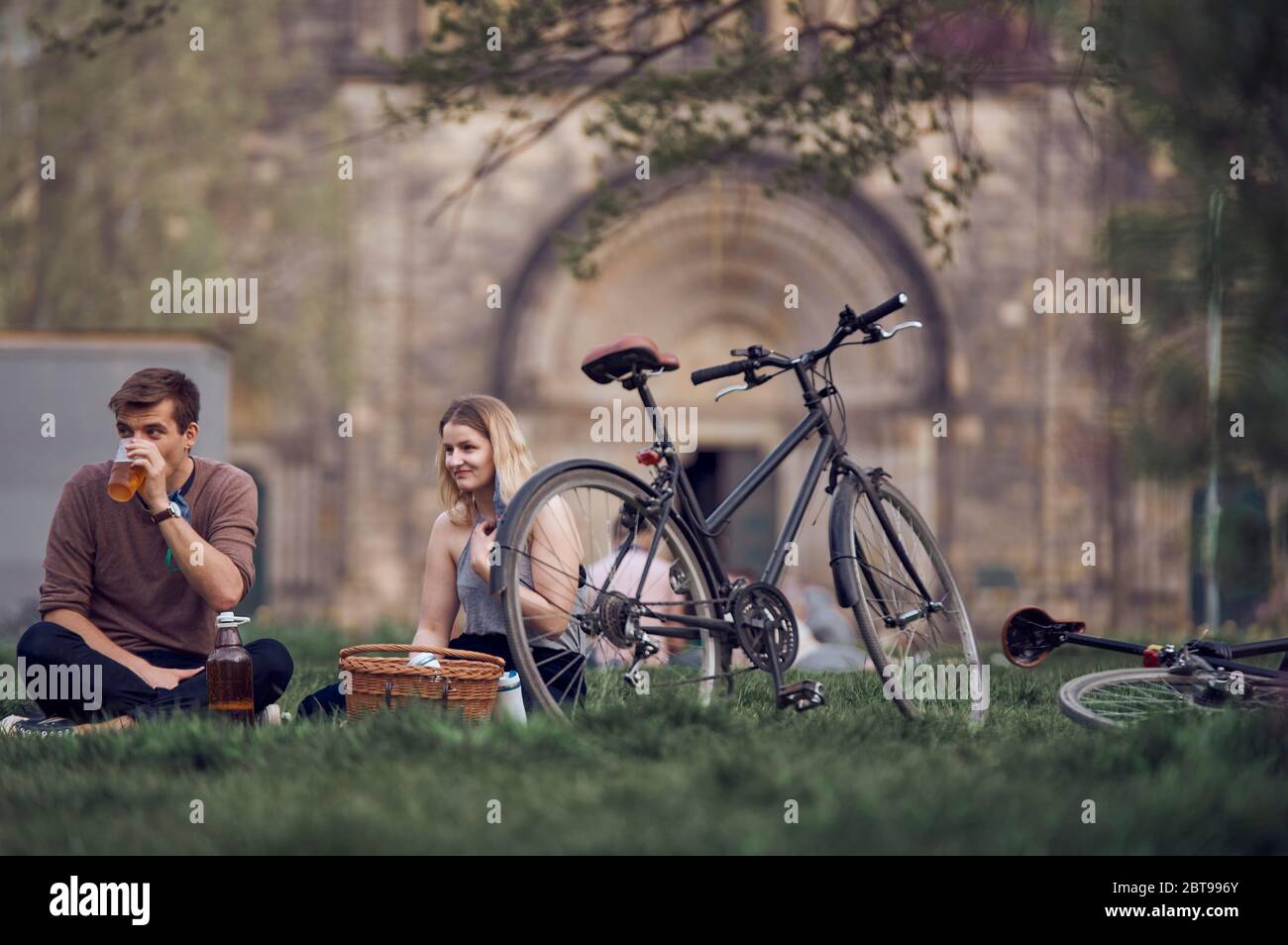 Ein weißes junges Paar beim Picknick in einem Park mit Vintage-Fahrrädern, Picknickkorb und Bier aus Plastikkappe Stockfoto