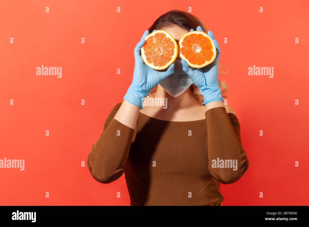 Frau in Schutzhandschuhen und Gesichtsmaske, die Augen mit orangefarbenen frischen Früchten, natürlichen Vitaminen für das Immunsystem und Grippe-Behandlung, gesunde Ernährung Stockfoto