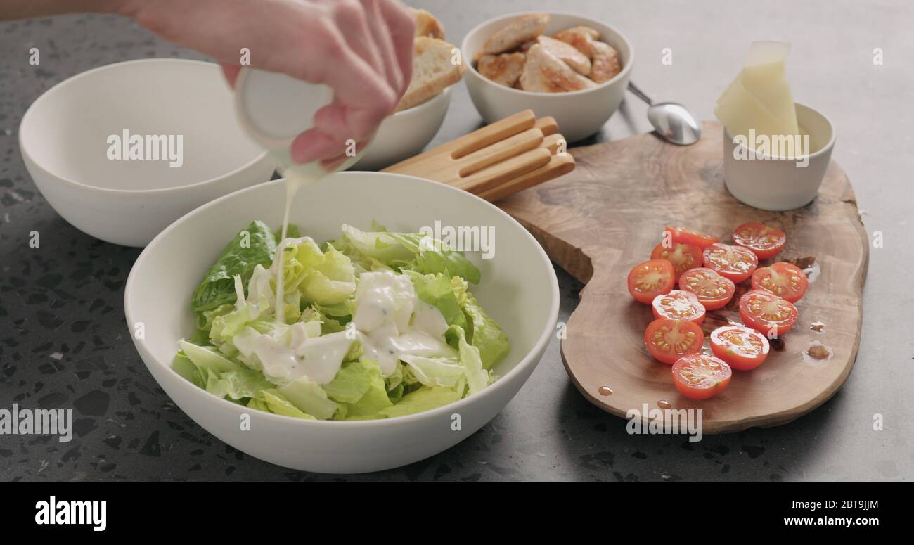 Man Hand fügen Sie Romaine Salat und Dressing caesar zu machen Salat in weißer Keramikschale auf Betonplatte Stockfoto