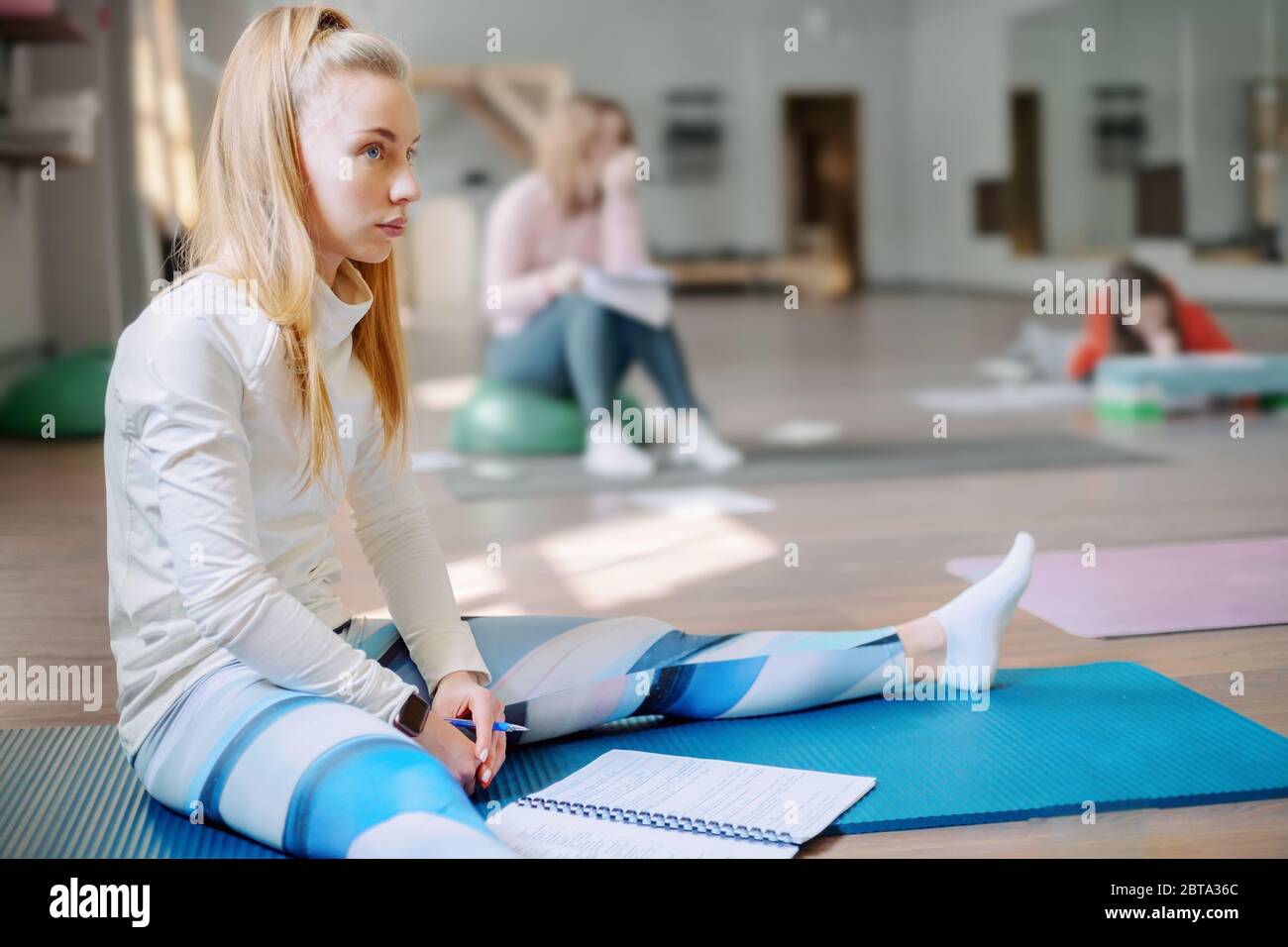 Schöne Blondine mit lockeren Haaren sitzt in einem Fitnessstudio und hört sich die Meisterklasse genau an. Pilates Lernen Stockfoto