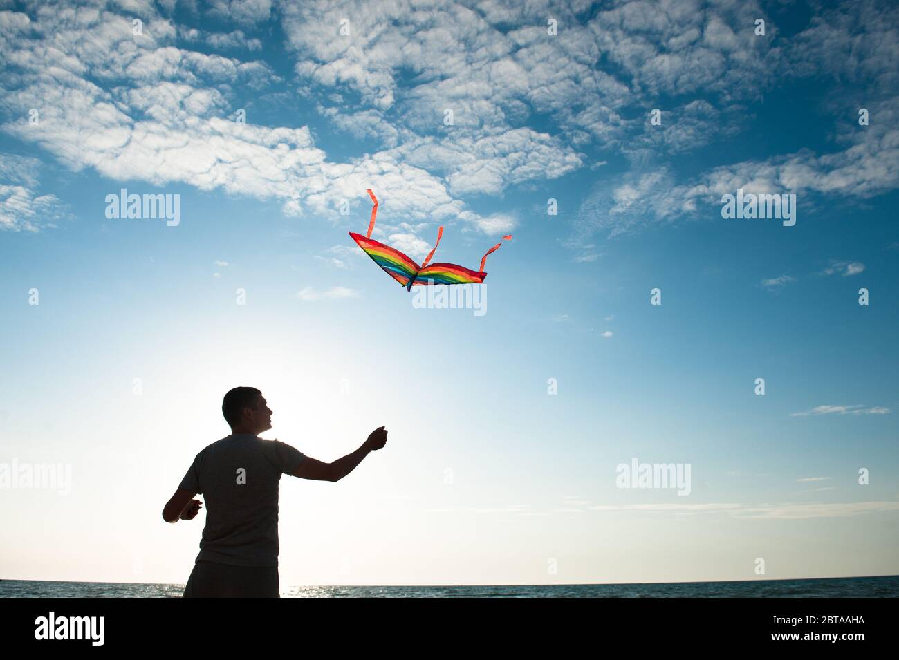 Junger aktiver Mann mit fliegenden bunten Drachen auf dem Meer und blauen Sonnenuntergang Himmel Hintergrund Stockfoto