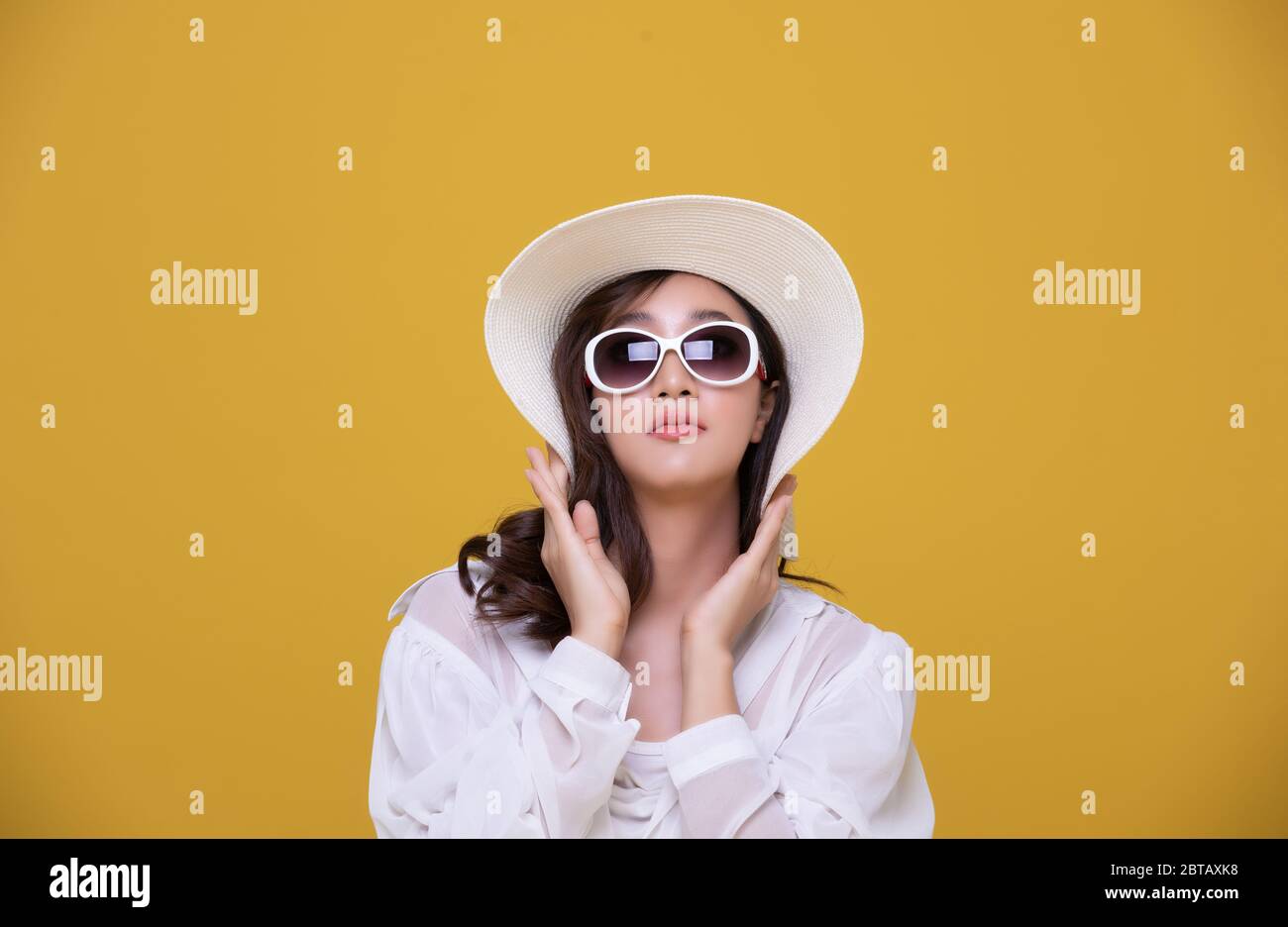 Portrait Asian schöne glückliche junge Frau mit Sonnenbrille und Hut lächelnd fröhlich im Sommer und Blick auf Kamera isoliert auf gelben Studio backgrou Stockfoto