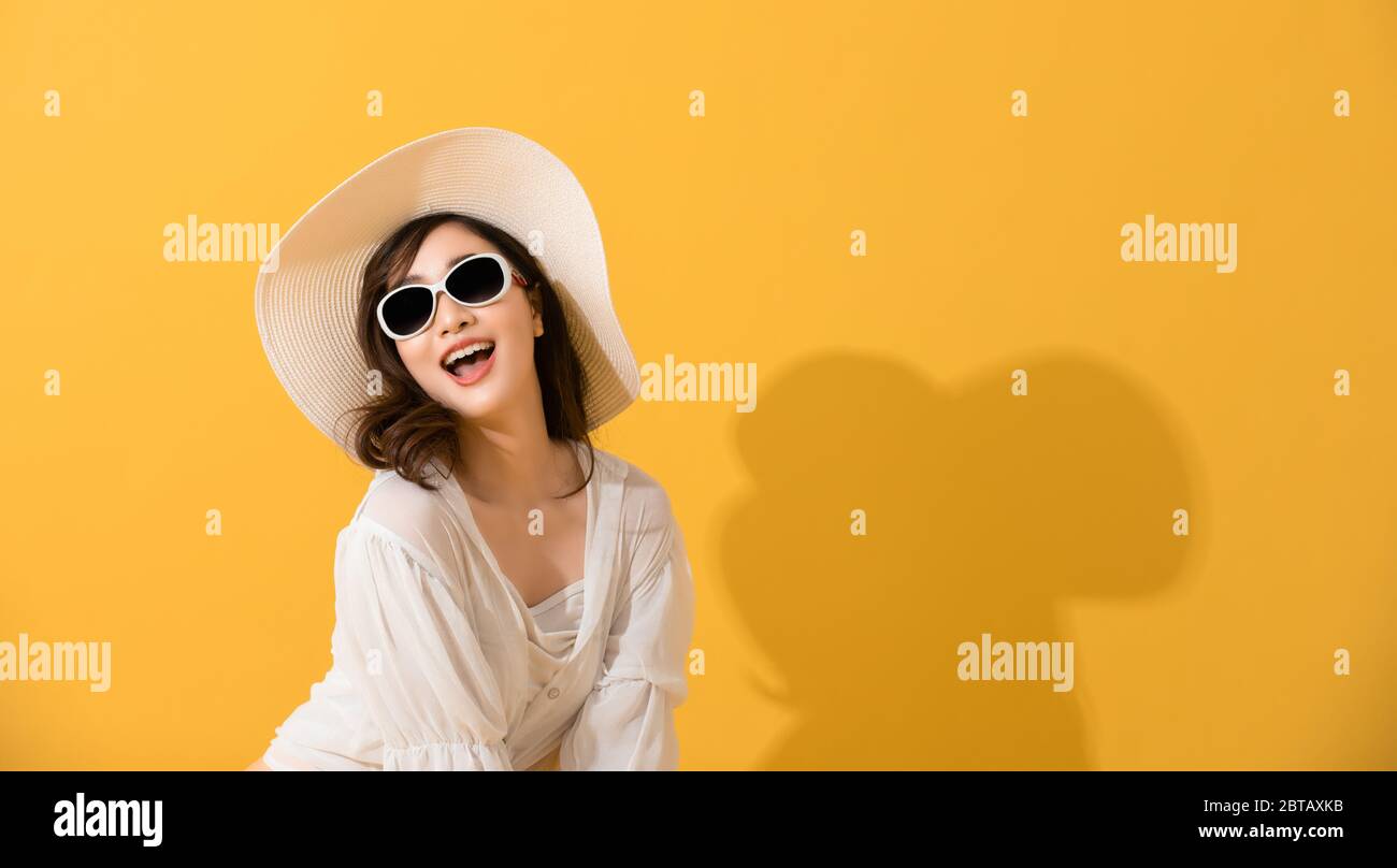 Portrait Asian schöne glückliche junge Frau mit Sonnenbrille und Hut lächelnd fröhlich im Sommer und Blick auf Kamera isoliert auf gelben Studio backgrou Stockfoto