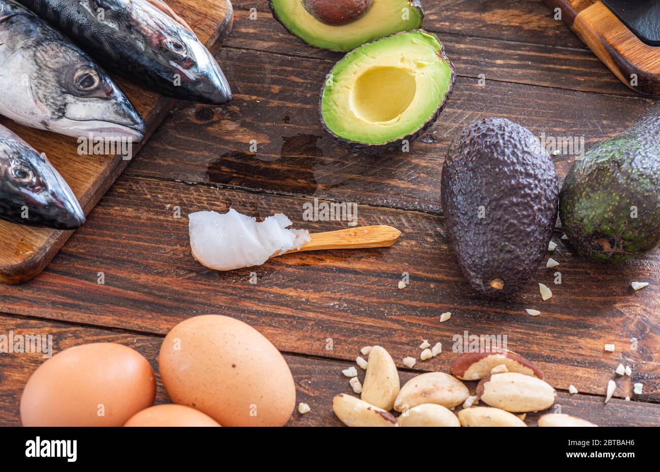 Nahaufnahme von ketogenem Futter: MCT-Öl, Makrelenfisch, Avocado, Eier, Kokosbutter, brasilianische Nüsse, Radieschen, Omega-3-Ergänzungen auf einem Holztisch Stockfoto