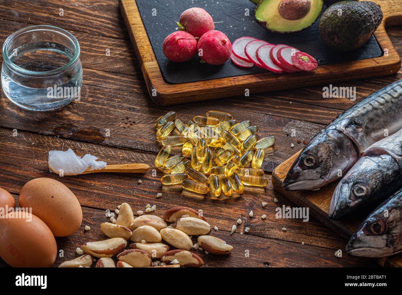 Nahaufnahme von ketogenem Futter: MCT-Öl, Makrelenfisch, Avocado, Eier, Kokosbutter, brasilianische Nüsse, Radieschen, Omega-3-Ergänzungen auf einem Holztisch Stockfoto