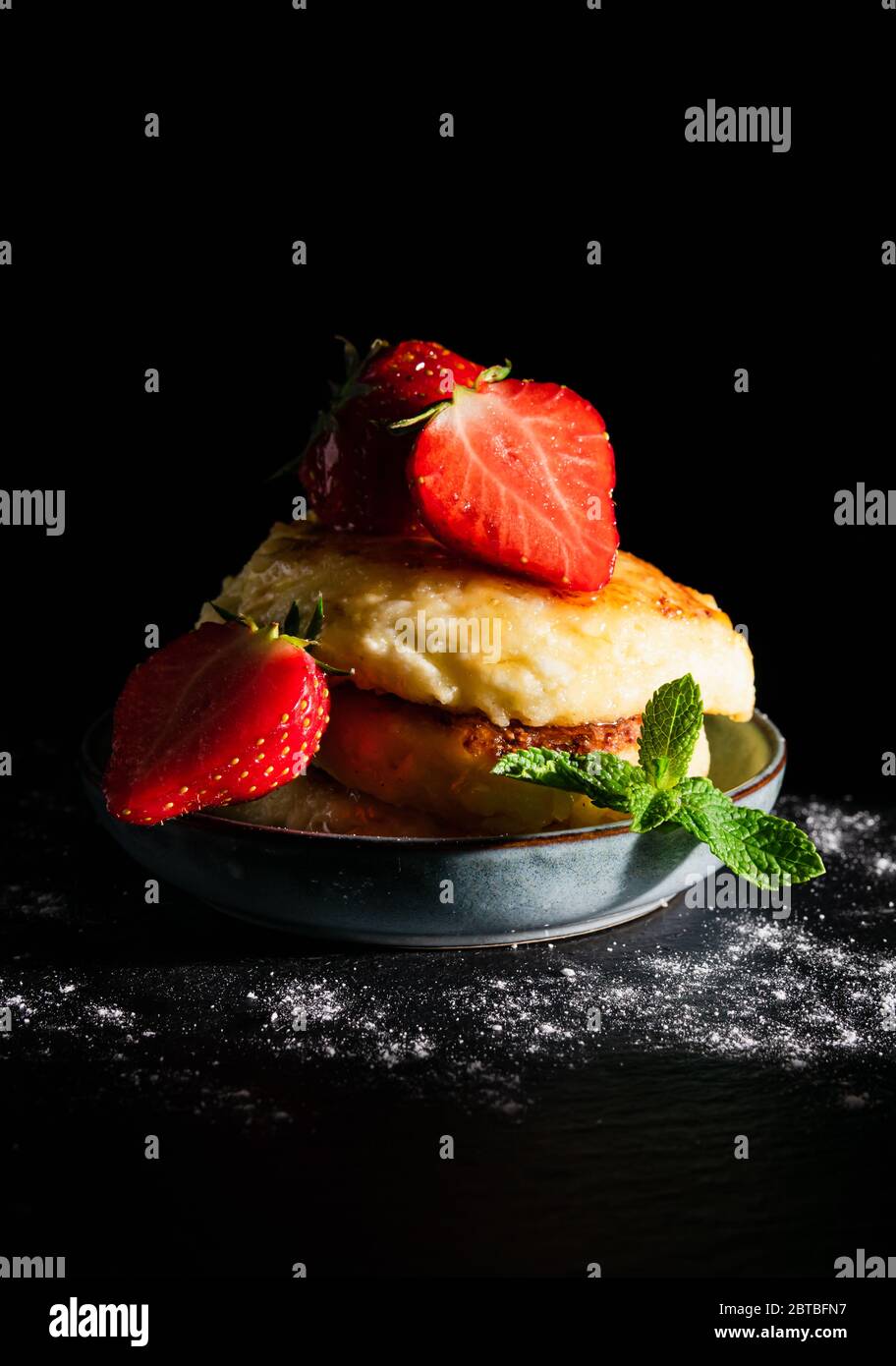 Stillleben Käsekuchen mit Erdbeeren und einem Blatt Minze, mit Honig auf schwarzem Hintergrund bestreut Stockfoto