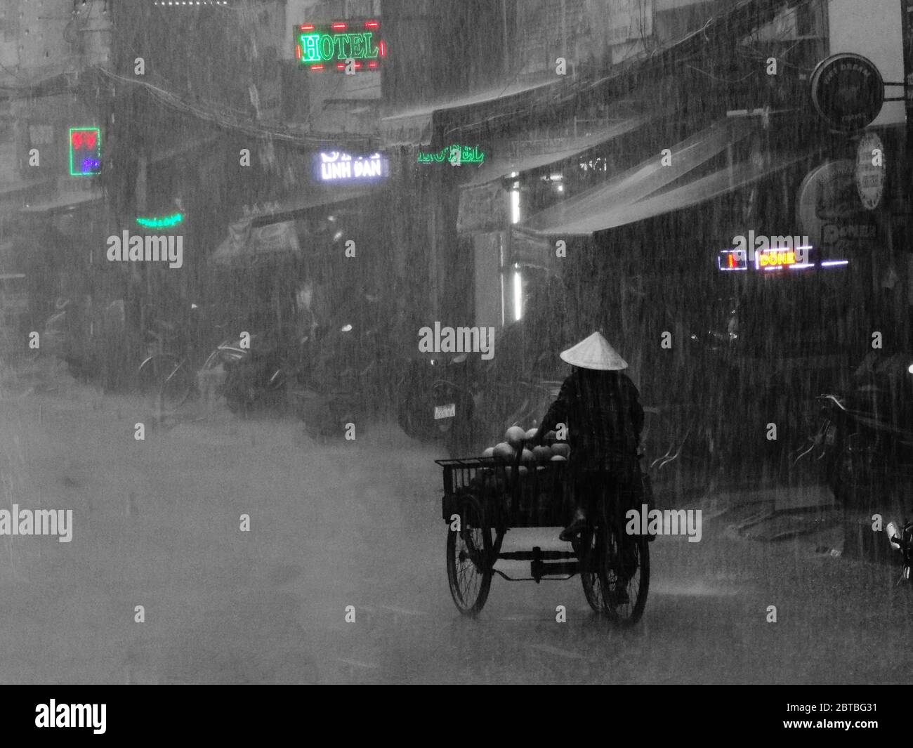 Vietnamesischer Mann, der Lebensmittel mit dem Fahrrad unter dem Regen in Ho Chi Minh Stadt in Vietnam transportiert Stockfoto