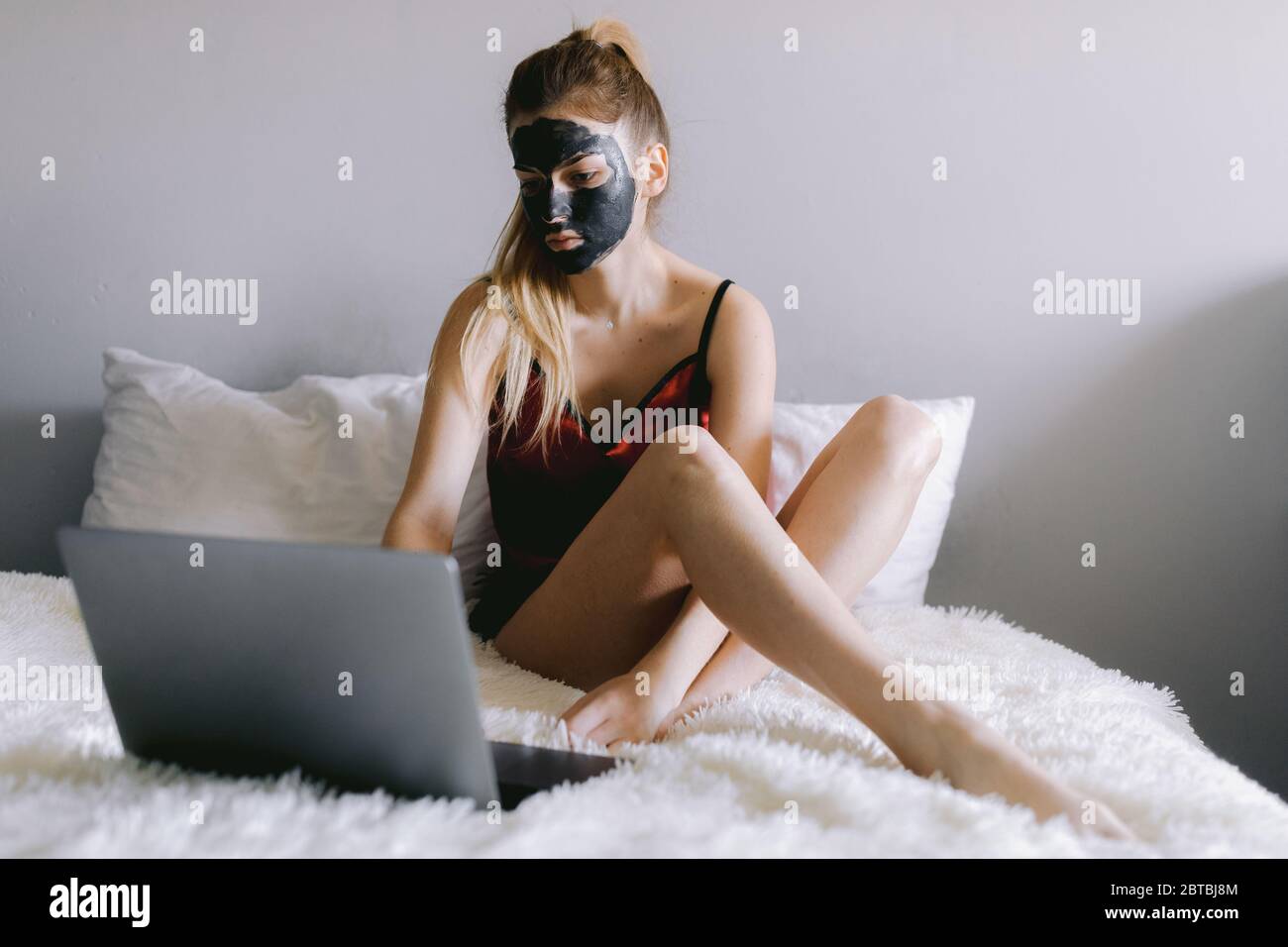 Junge Frau in Pyjama und mit Lehmmaske auf Gesicht sitzen auf dem Bett und  Video auf Laptop während Spa-Routine zu Hause Stockfotografie - Alamy