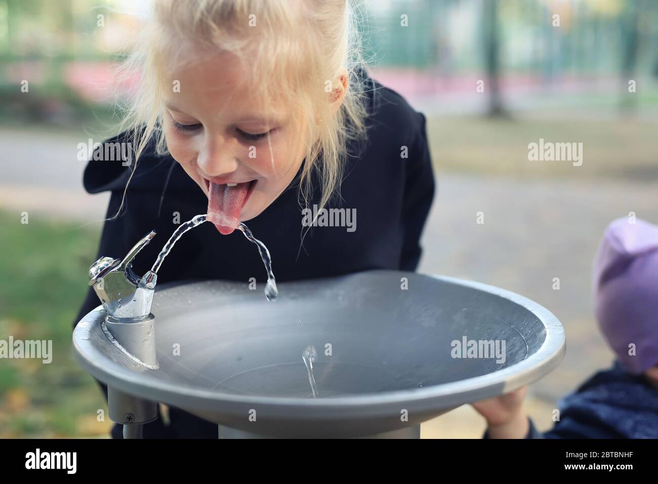 Nette liebenswert kaukasischen blonde kleine durstige Schulmädchen Trinkwasser aus öffentlichen trinkbaren Brunnen Wasserhahn im Stadtpark auf hellen heißen Sommertag Stockfoto