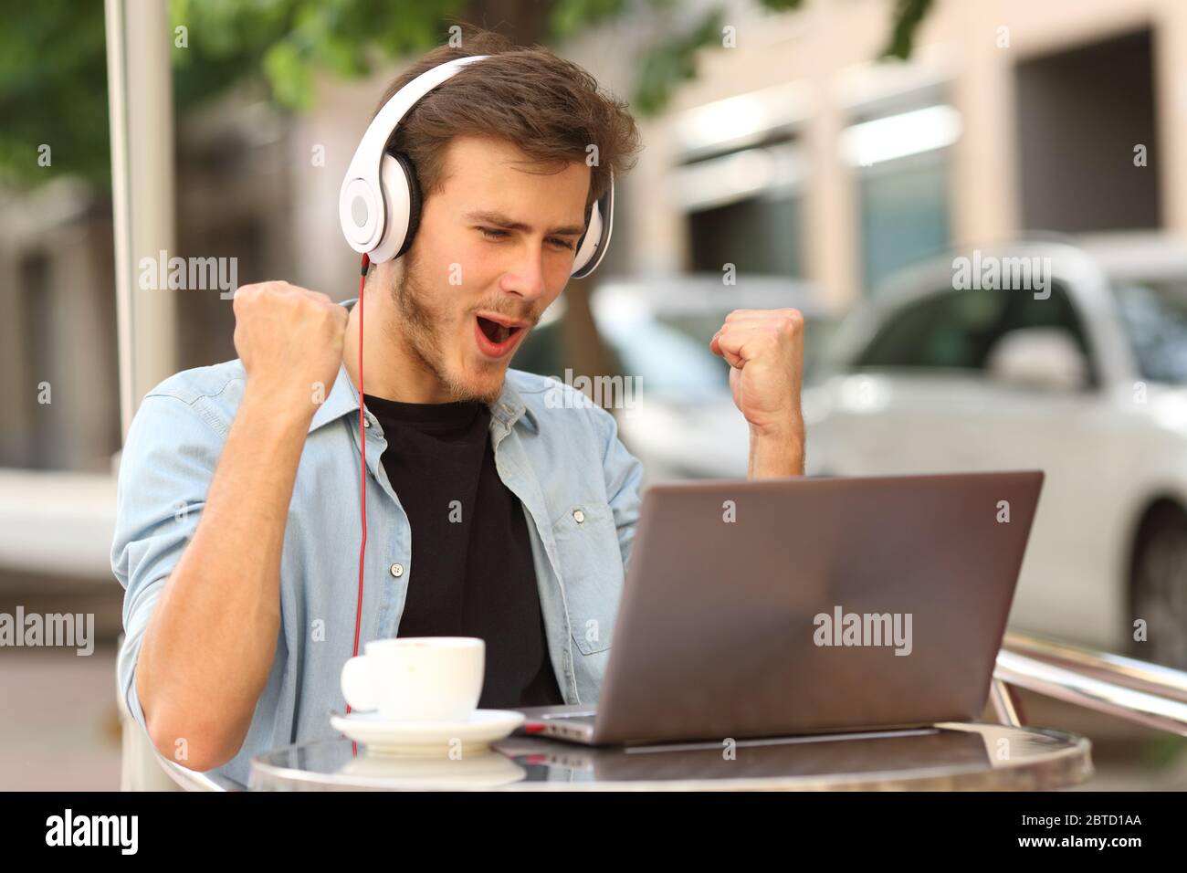 Aufgeregt Mann mit Kopfhörer feiern gute Nachrichten auf Laptop sitzen auf einer Bar Terrasse Stockfoto