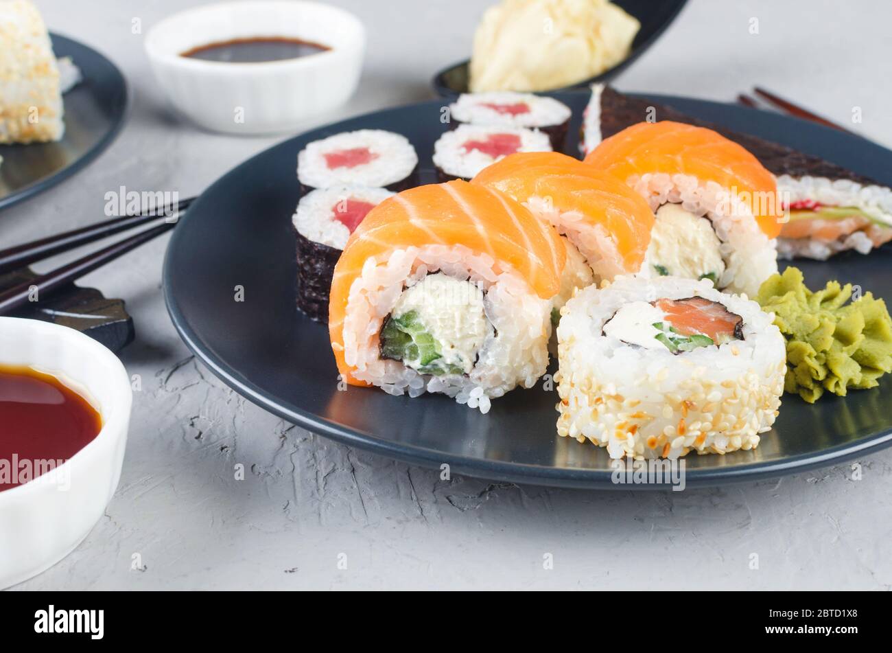 Verschiedene Sushi-Brötchen auf einem Teller und Sojasoße, Ingwer, Wasabi und Essstäbchen auf einem grauen Betontisch. Asiatische Speisekarte. Lieferservice Stockfoto