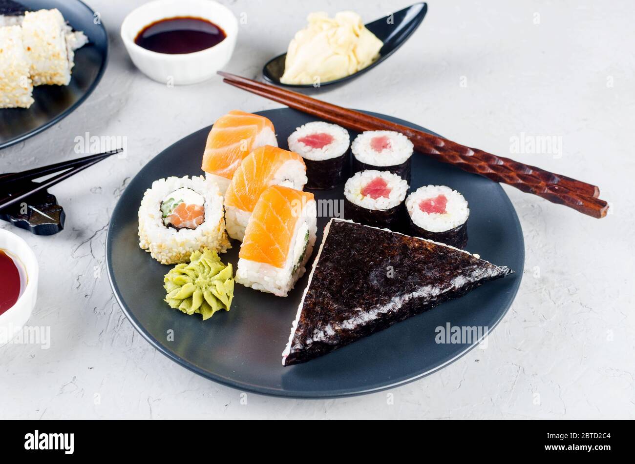 Verschiedene Sushi-Brötchen auf einem Teller und Sojasoße, Ingwer, Wasabi und Essstäbchen auf einem grauen Betontisch. Asiatische Speisekarte. Lieferservice Stockfoto
