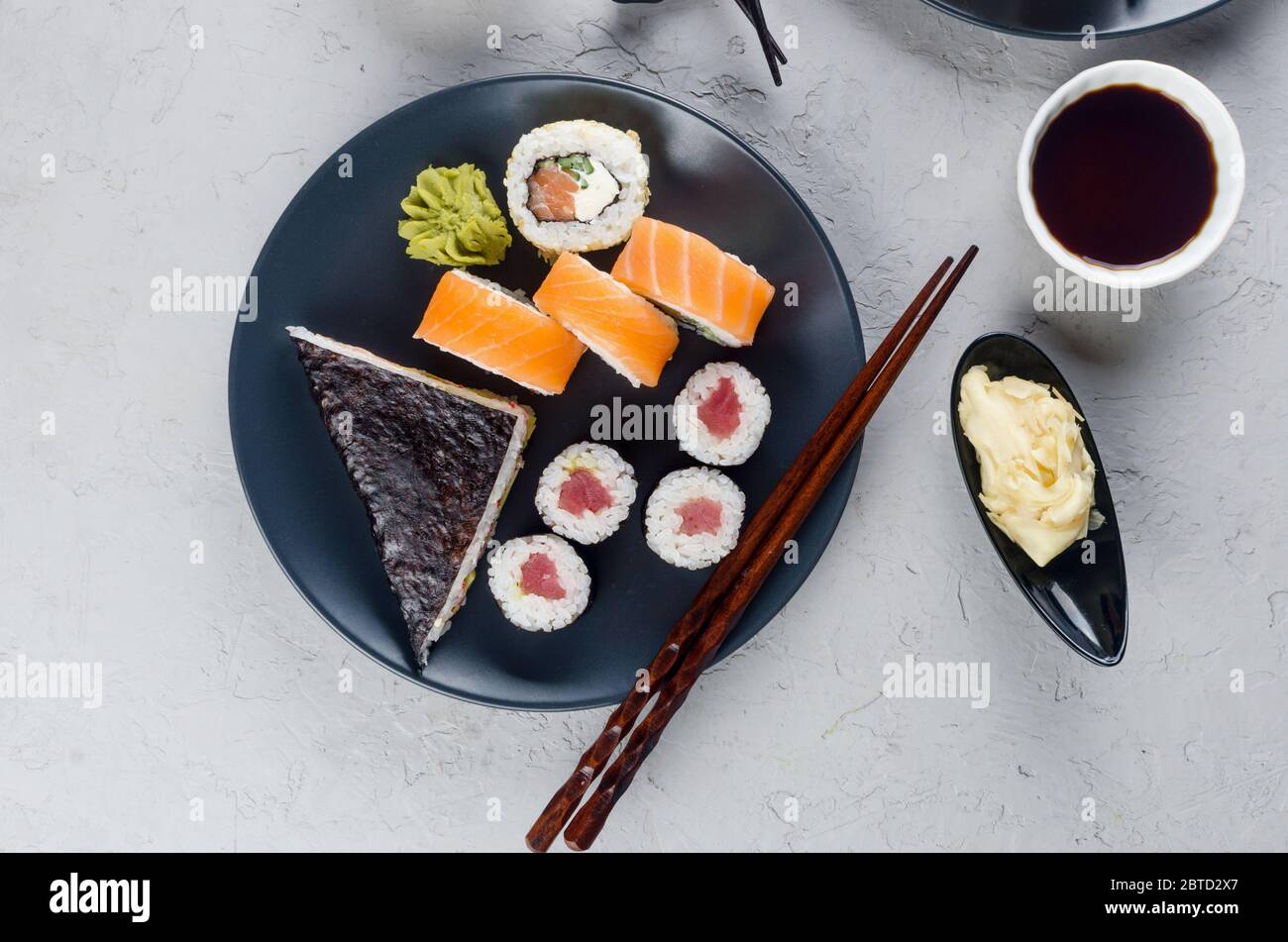 Verschiedene Sushi-Brötchen auf einem Teller und Sojasoße, Ingwer, Wasabi und Essstäbchen auf einem grauen Betontisch. Asiatische Speisekarte. Lieferservice Stockfoto