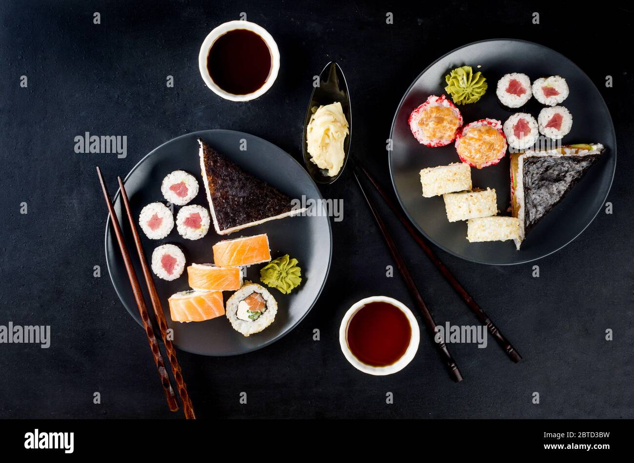 Verschiedene Sushi-Brötchen auf einem Teller und Sojasoße, Ingwer, Wasabi und Essstäbchen auf einem schwarzen Tisch. Asiatisches Restaurant, Sushi-Menü. Lieferservice Stockfoto