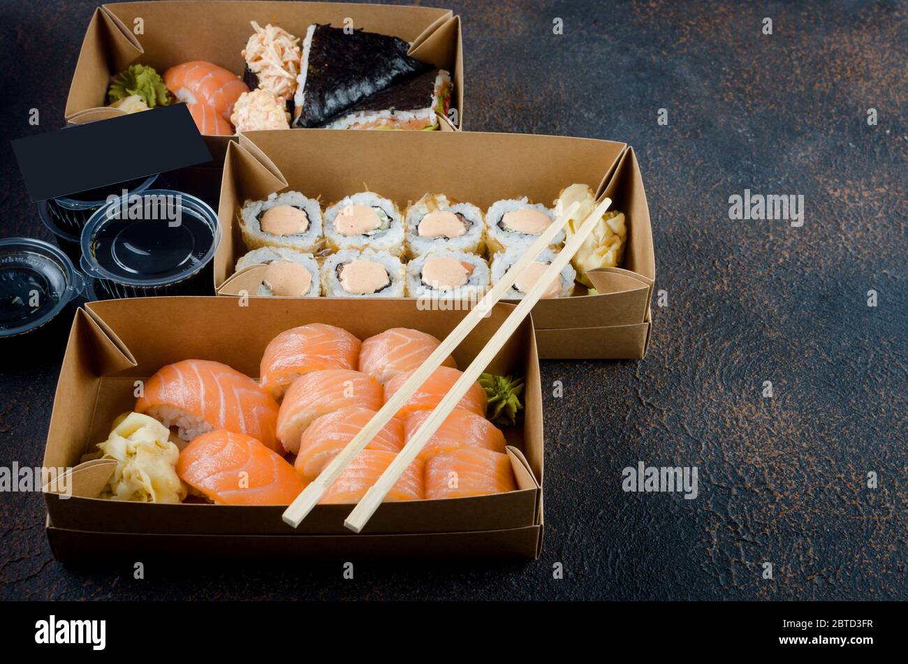 Leckere Sushi-Rollen in Einweg-Kraftpapier-Boxen, Saucen auf dunklem Tisch. Konzept Lieferservice Japan Lebensmittel in Öko-Container. Flach legen, Vorlage Mock Stockfoto
