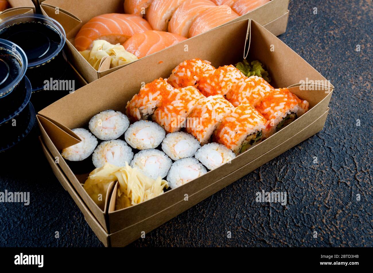 Leckere Sushi-Rollen in Einweg-Kraftpapier-Boxen, Saucen auf dunklem Tisch. Konzept Lieferservice Japan Lebensmittel in Öko-Container. Flach legen, Vorlage Mock Stockfoto
