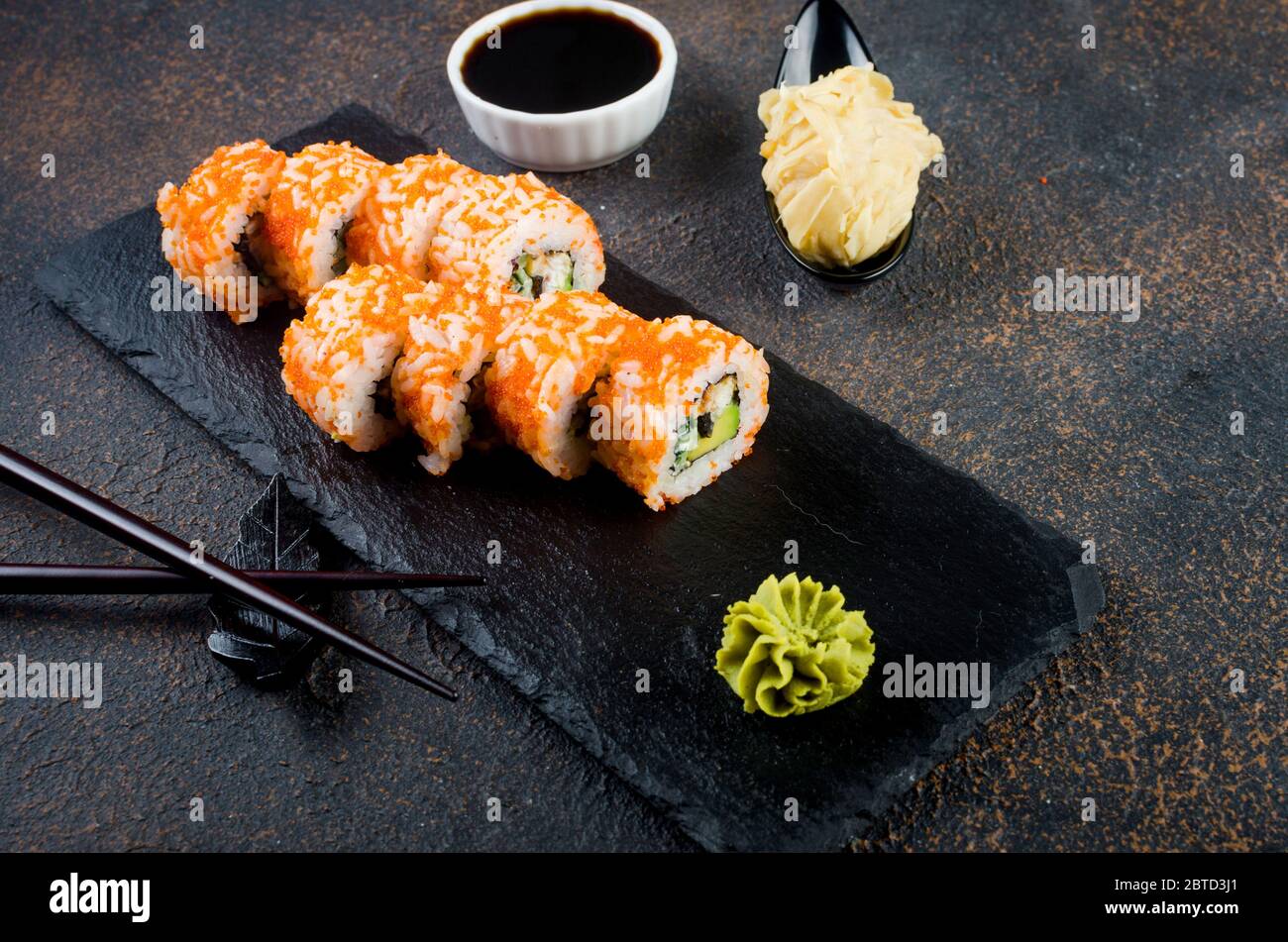Leckere kalifornische Brötchen und Maki auf Steinplatte mit Saucen, Essstäbchen, Ingwer und Wasabi auf dunklem Tisch. Asiatische Küche. Lieferservice Japanisch foo Stockfoto