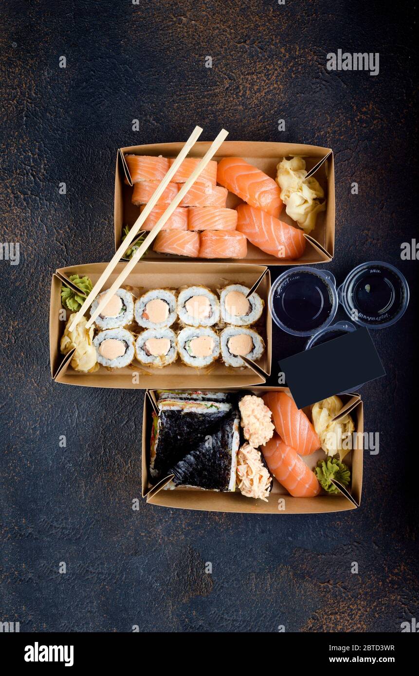 Leckere Sushi-Rollen in Einweg-Kraftpapier-Boxen, Saucen auf dunklem Tisch. Konzept Lieferservice Japan Lebensmittel in Öko-Container. Flach legen, Vorlage Mock Stockfoto