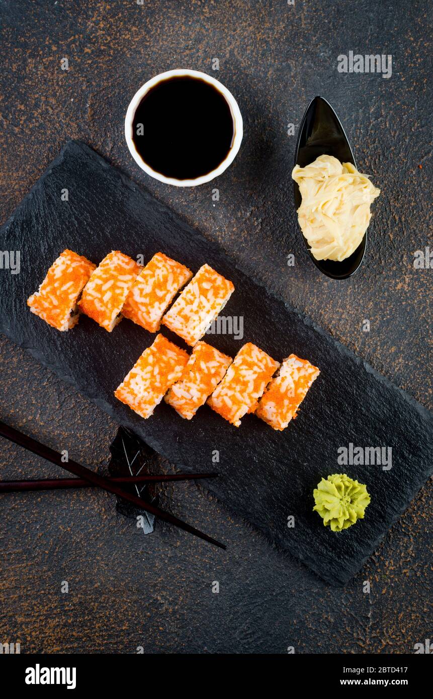 Leckere kalifornische Brötchen und Maki auf Steinplatte mit Saucen, Essstäbchen, Ingwer und Wasabi auf dunklem Tisch. Asiatische Küche. Lieferservice Japanisch foo Stockfoto