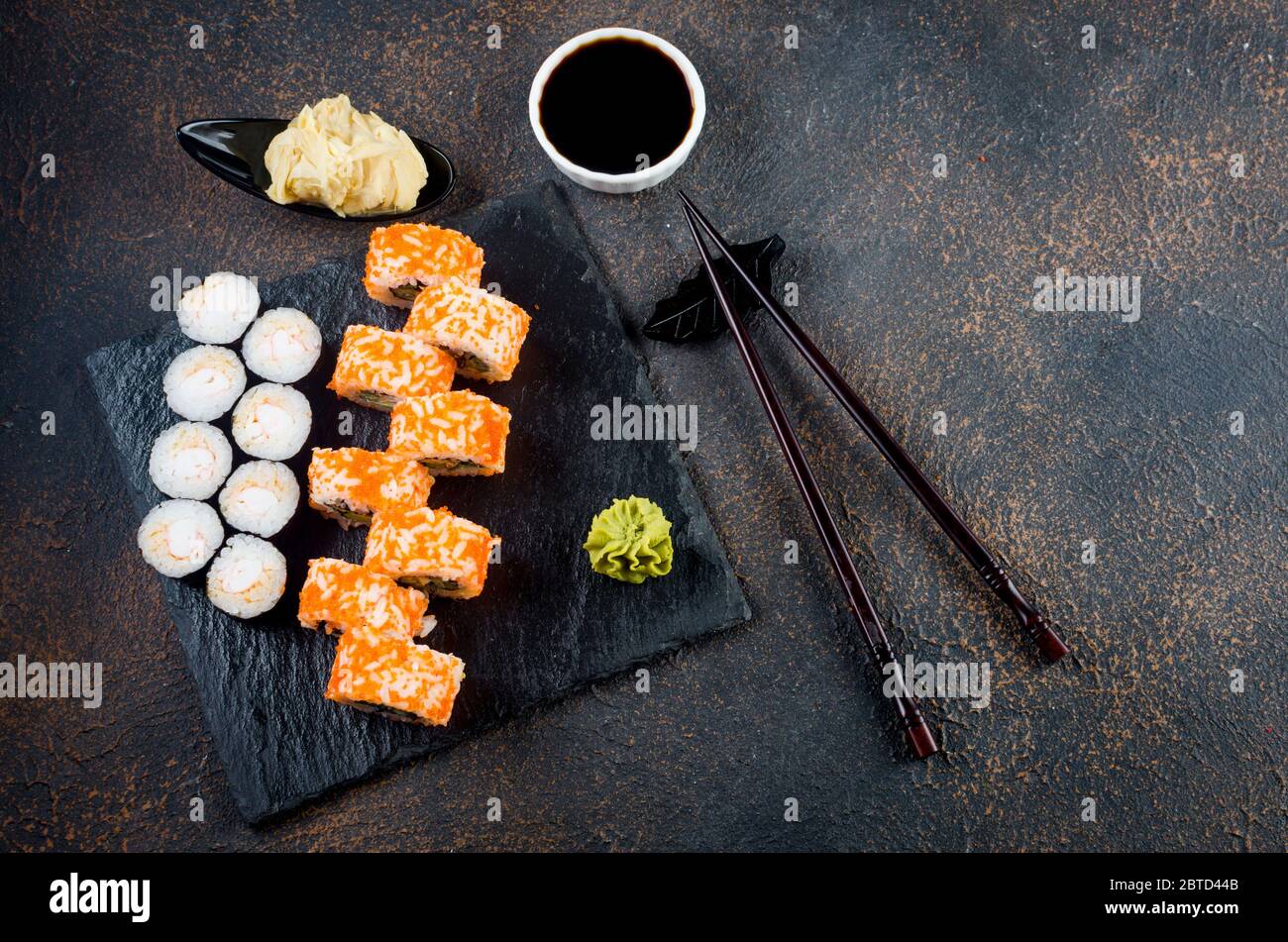 Leckere kalifornische Brötchen und Maki auf Steinplatte mit Saucen, Essstäbchen, Ingwer und Wasabi auf dunklem Tisch. Asiatische Küche. Lieferservice Japanisch foo Stockfoto