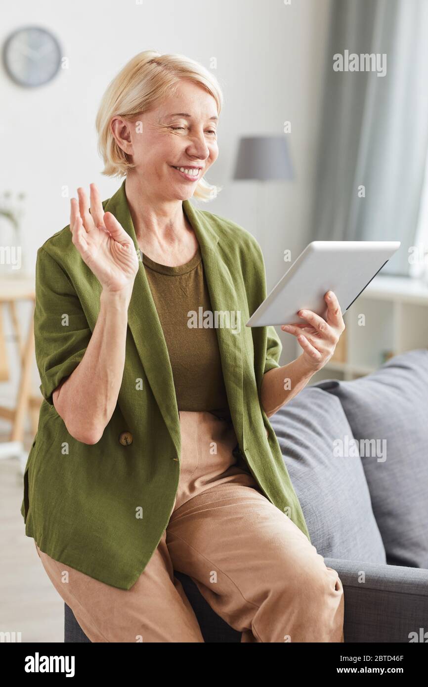 Ältere Frau in Freizeitkleidung, die auf ein digitales Tablet schaut und ihrem Freund lächelt, während er zu Hause sitzt Stockfoto