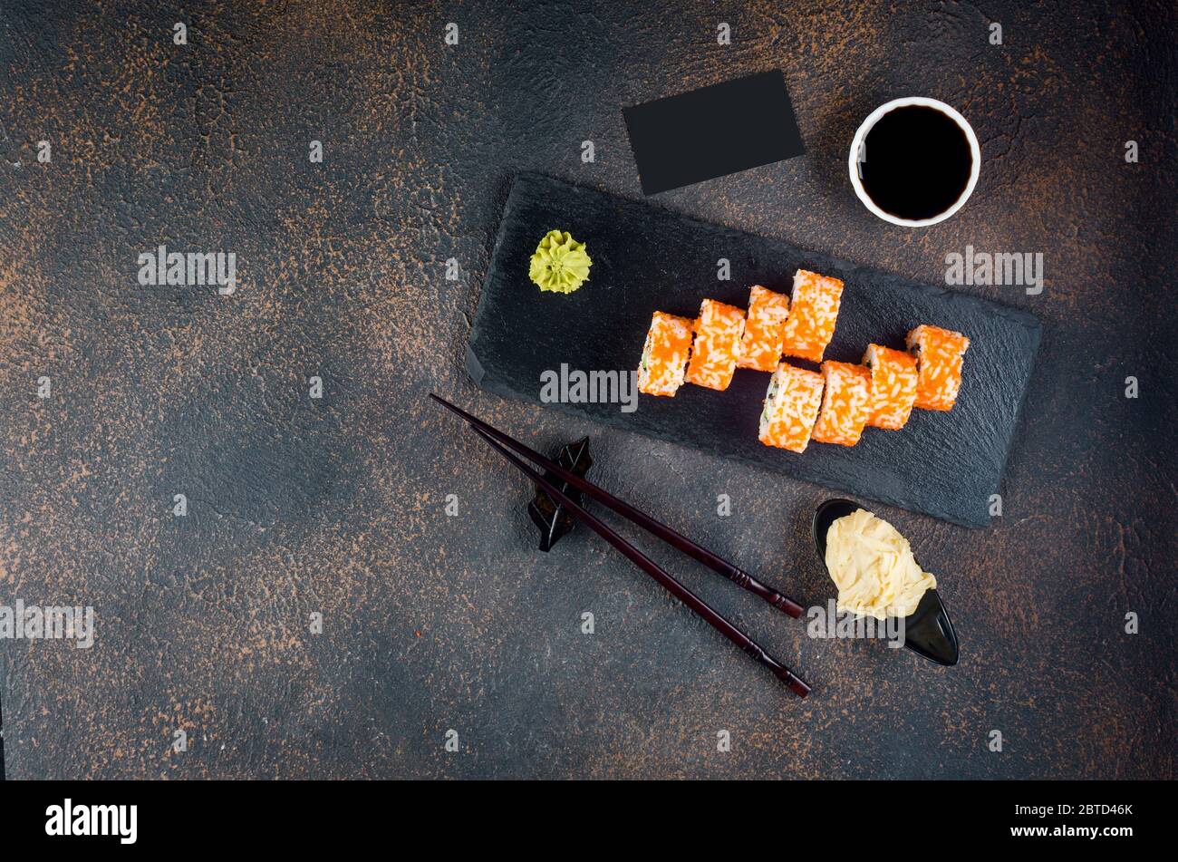 Leckere kalifornische Brötchen und Maki auf Steinplatte mit Saucen, Essstäbchen, Ingwer und Wasabi auf dunklem Tisch. Asiatische Küche. Lieferservice Japanisch foo Stockfoto