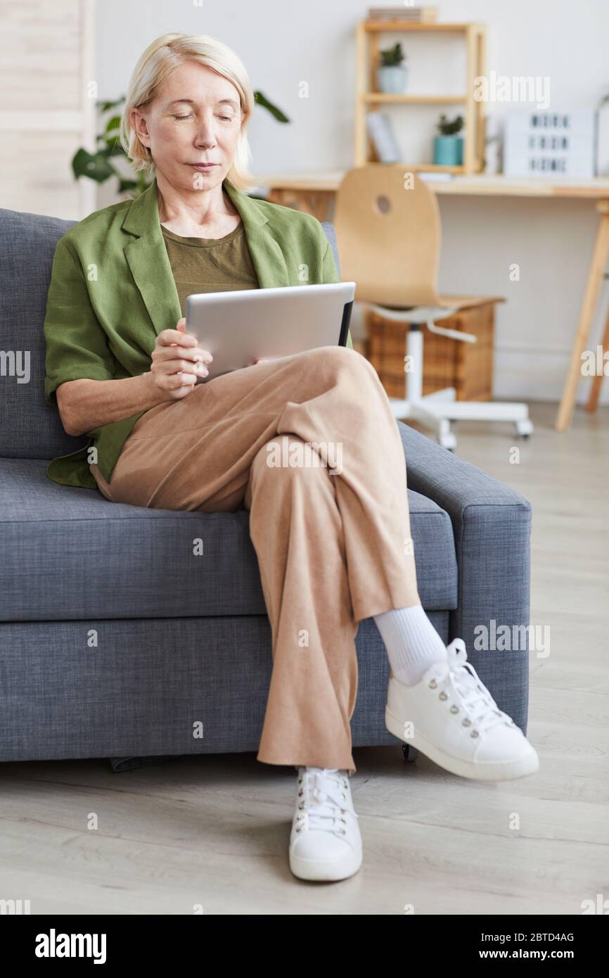 Reife Frau in Freizeitkleidung sitzt auf dem Sofa und sieht Filme online mit digitalen Tablet Stockfoto